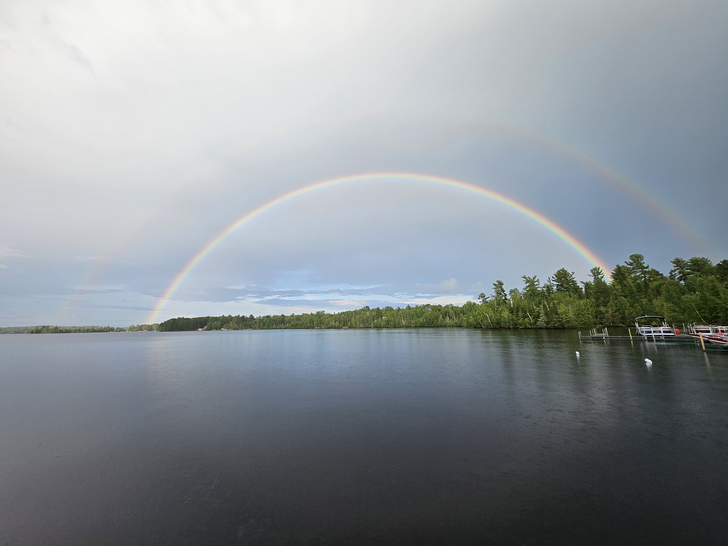 A calm lake with a clear double rainbow spanning across the sky and reflecting in the water. The horizon is lined with dense green trees, and the scene includes a dock with boats on the right side.