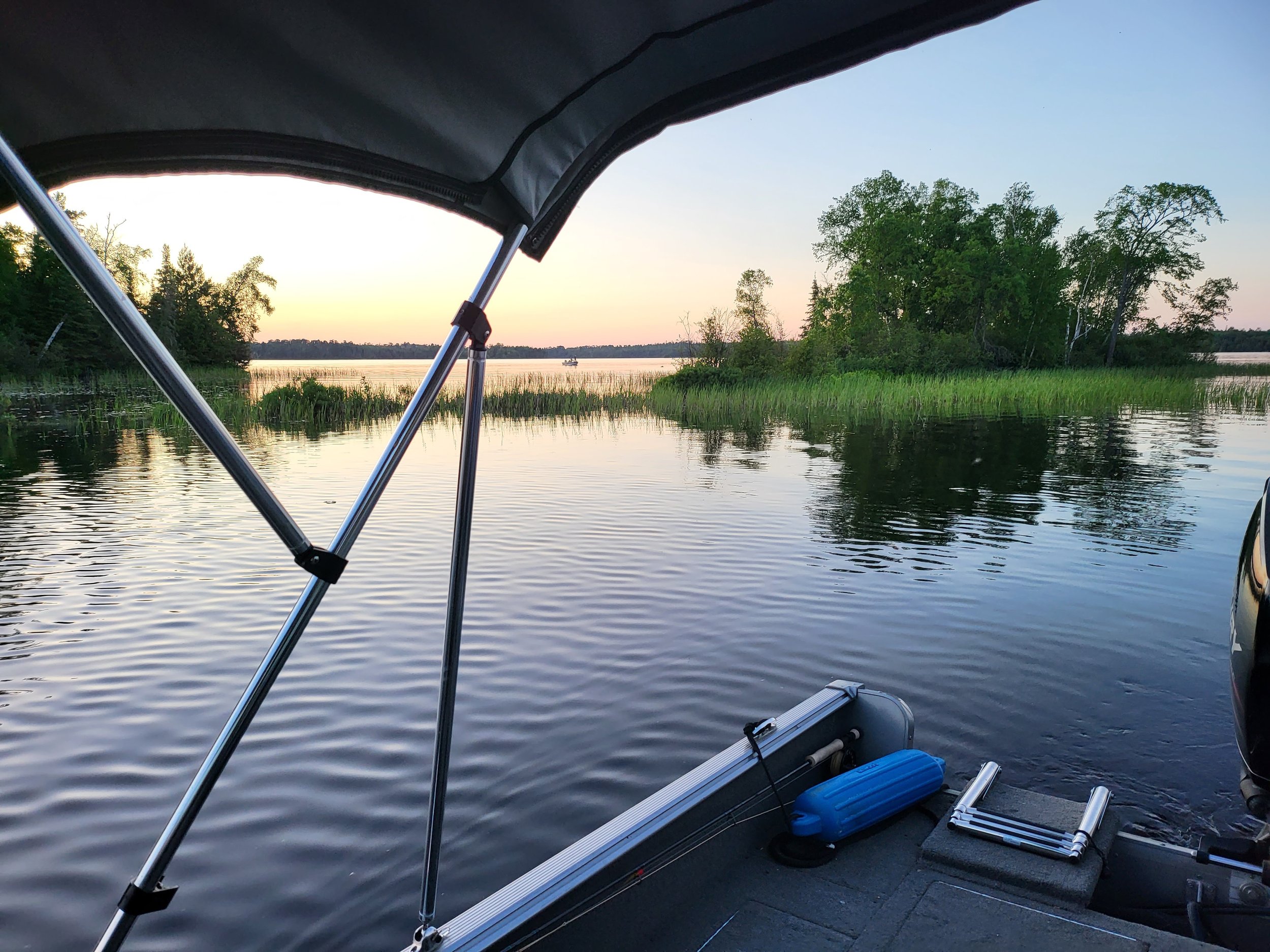 View from a boat on a lake at sunset, with calm water and a small island with trees and reeds visible in the distance. The boat canopy and a blue fender are visible in the foreground.