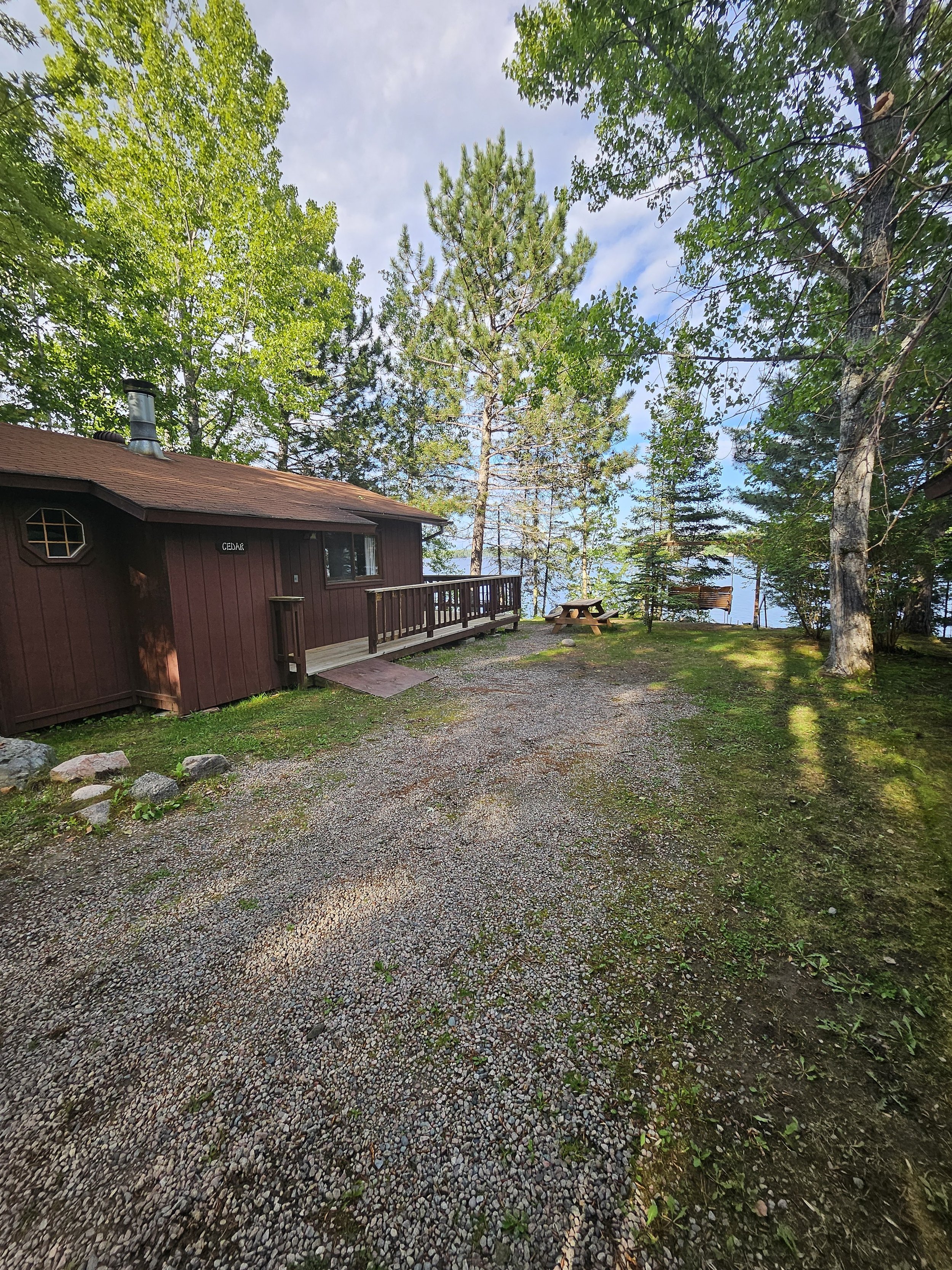 Rustic cabin amongst trees with a gravel path and a picnic table on a grassy area.