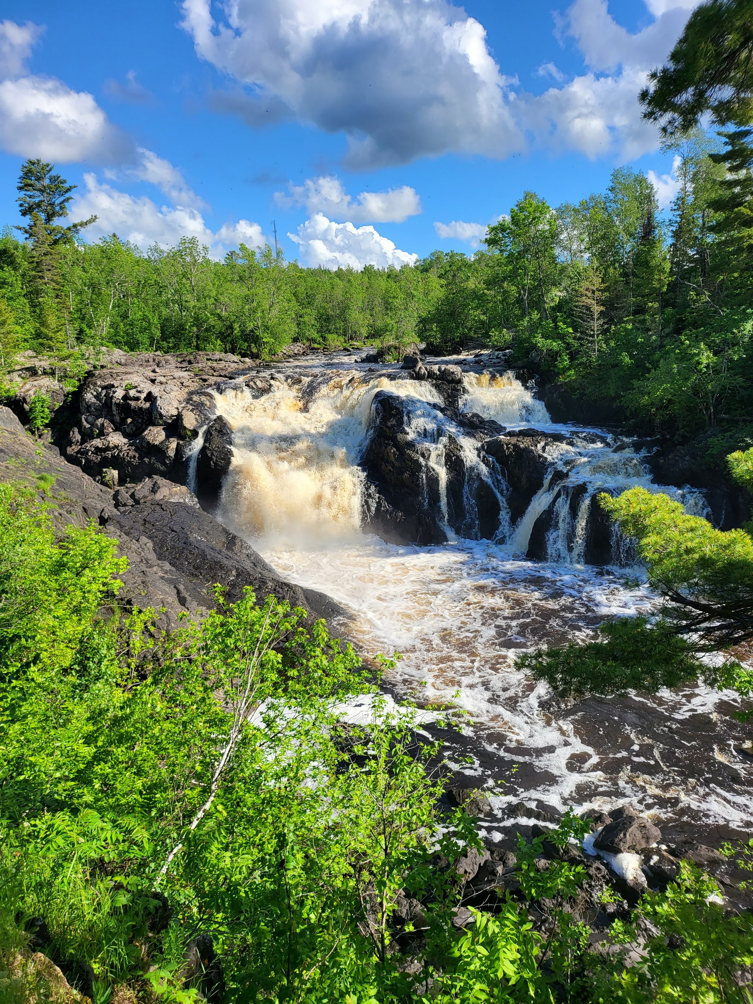 Waterfall cascading over rocks surrounded by lush green trees under a blue sky with clouds.