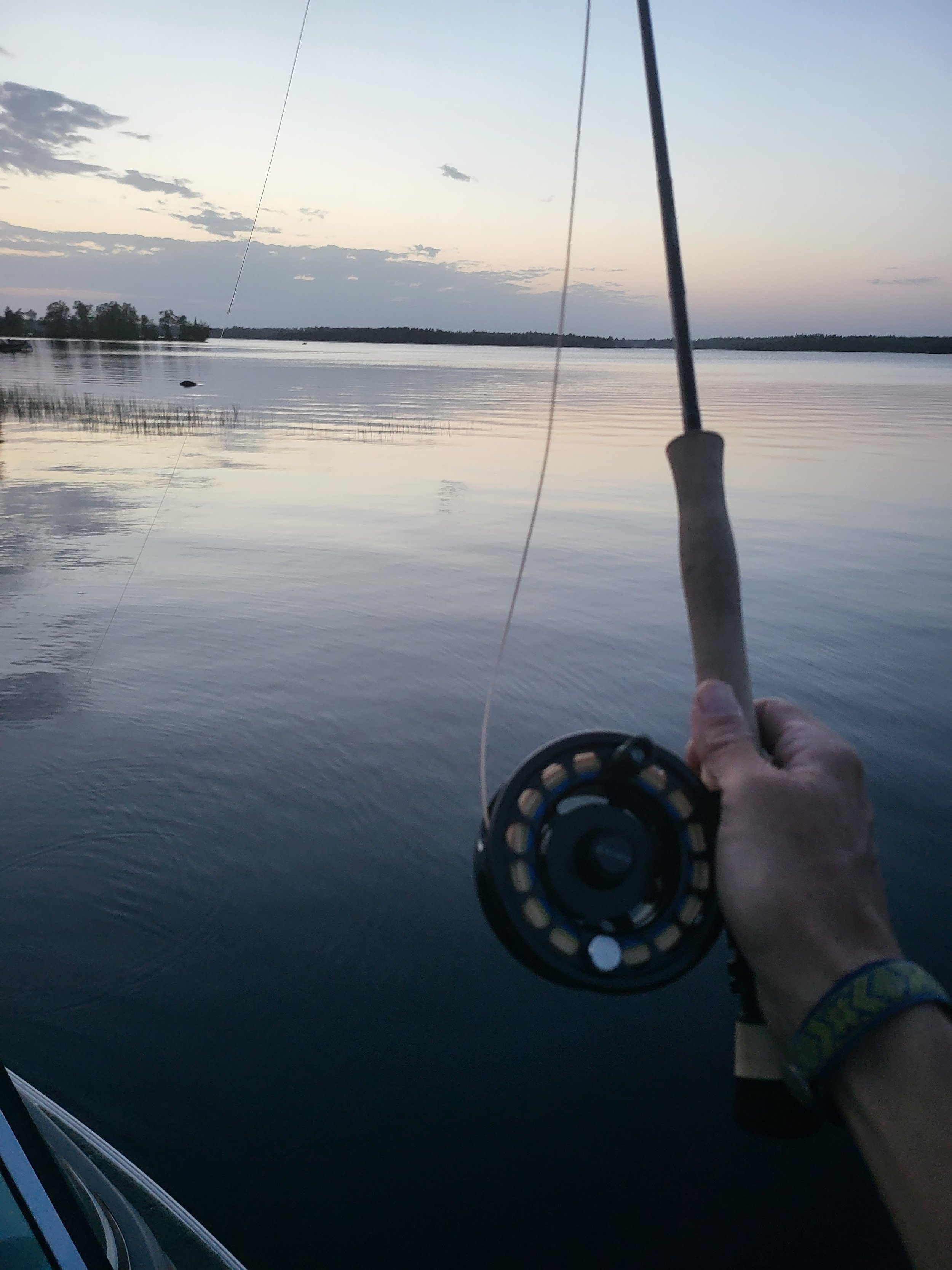 A person holding a fishing rod with a reel over a calm lake at sunset.