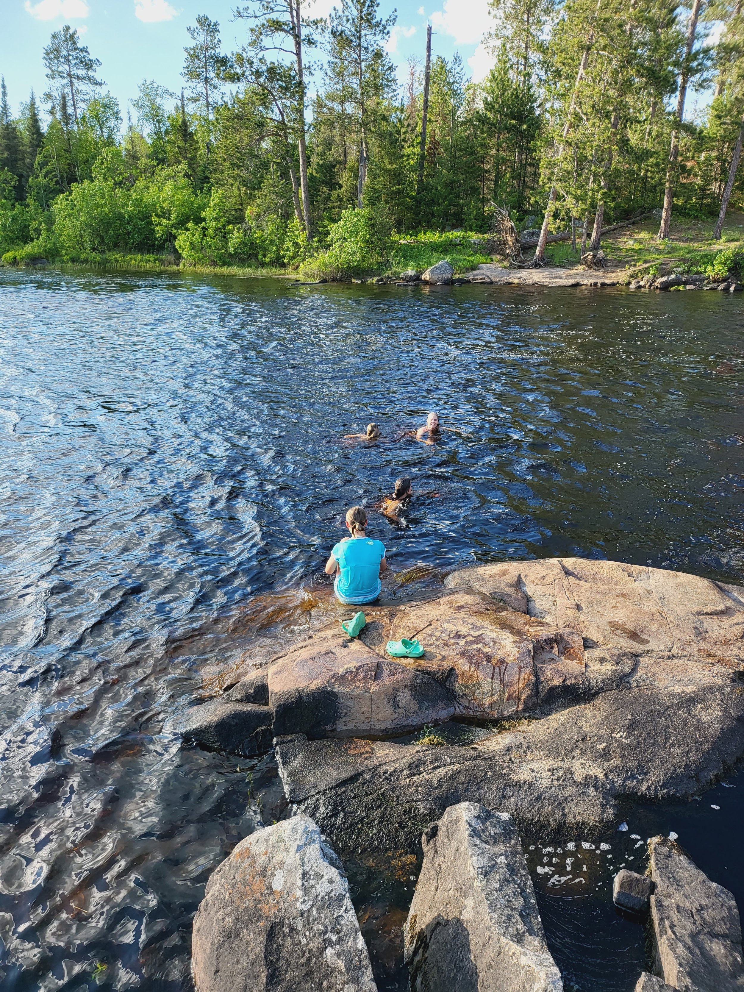 People enjoying swimming in a lake surrounded by pine trees, with one person sitting on a rock by the shore.