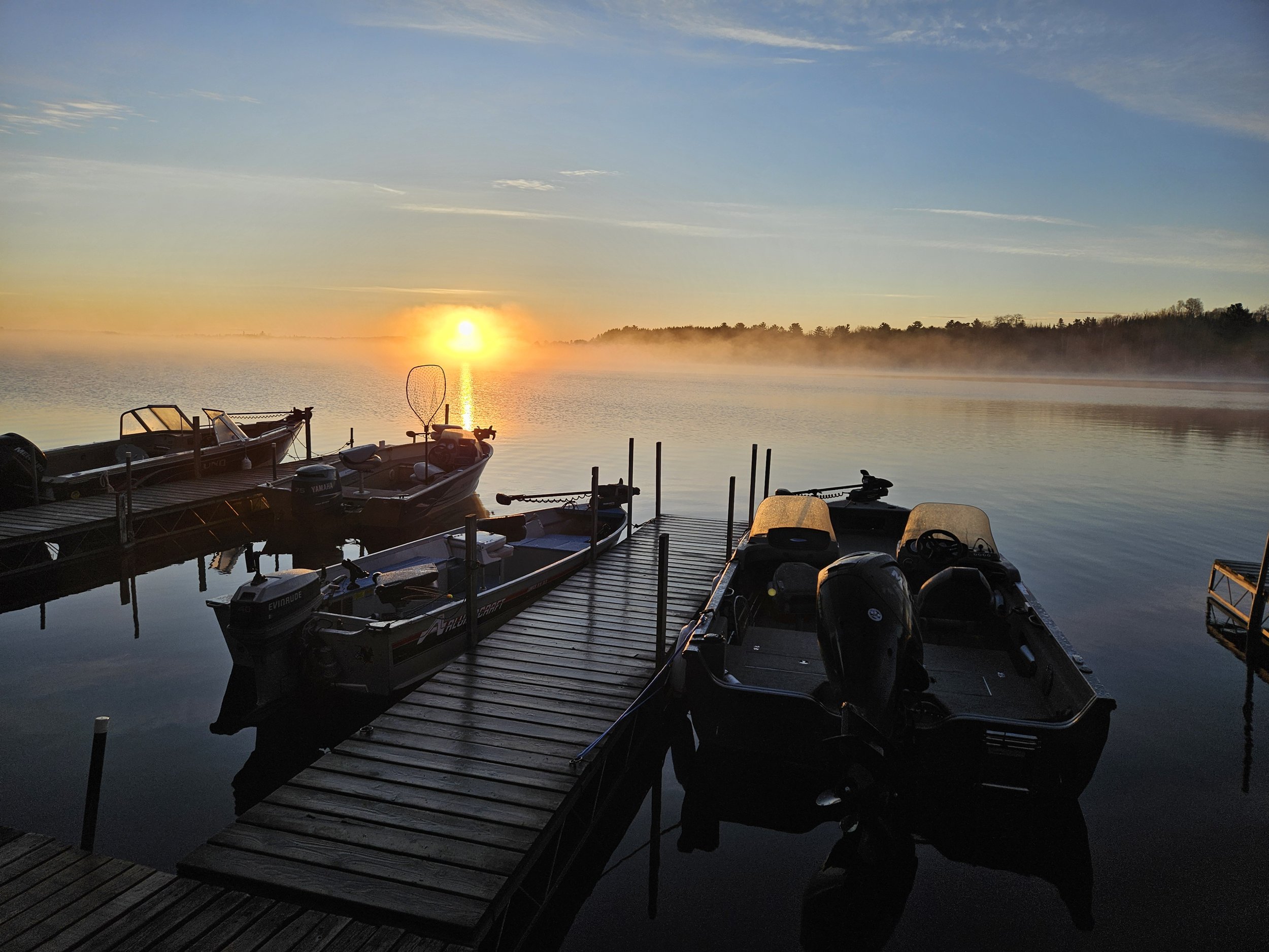 Boats docked at sunrise with mist over a calm lake.