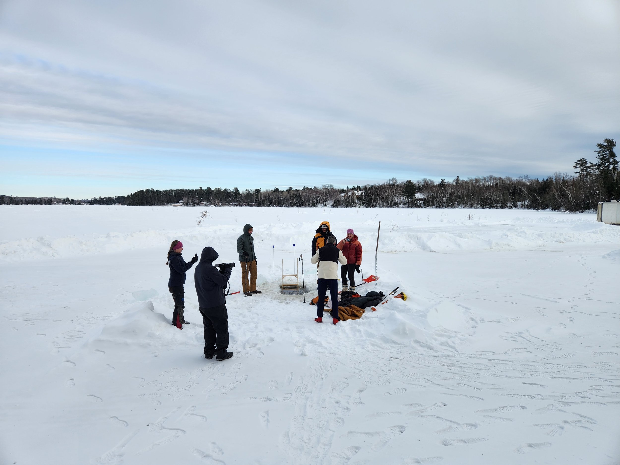 Group of people standing on a snowy landscape, with some engaging in ice fishing or winter activities.