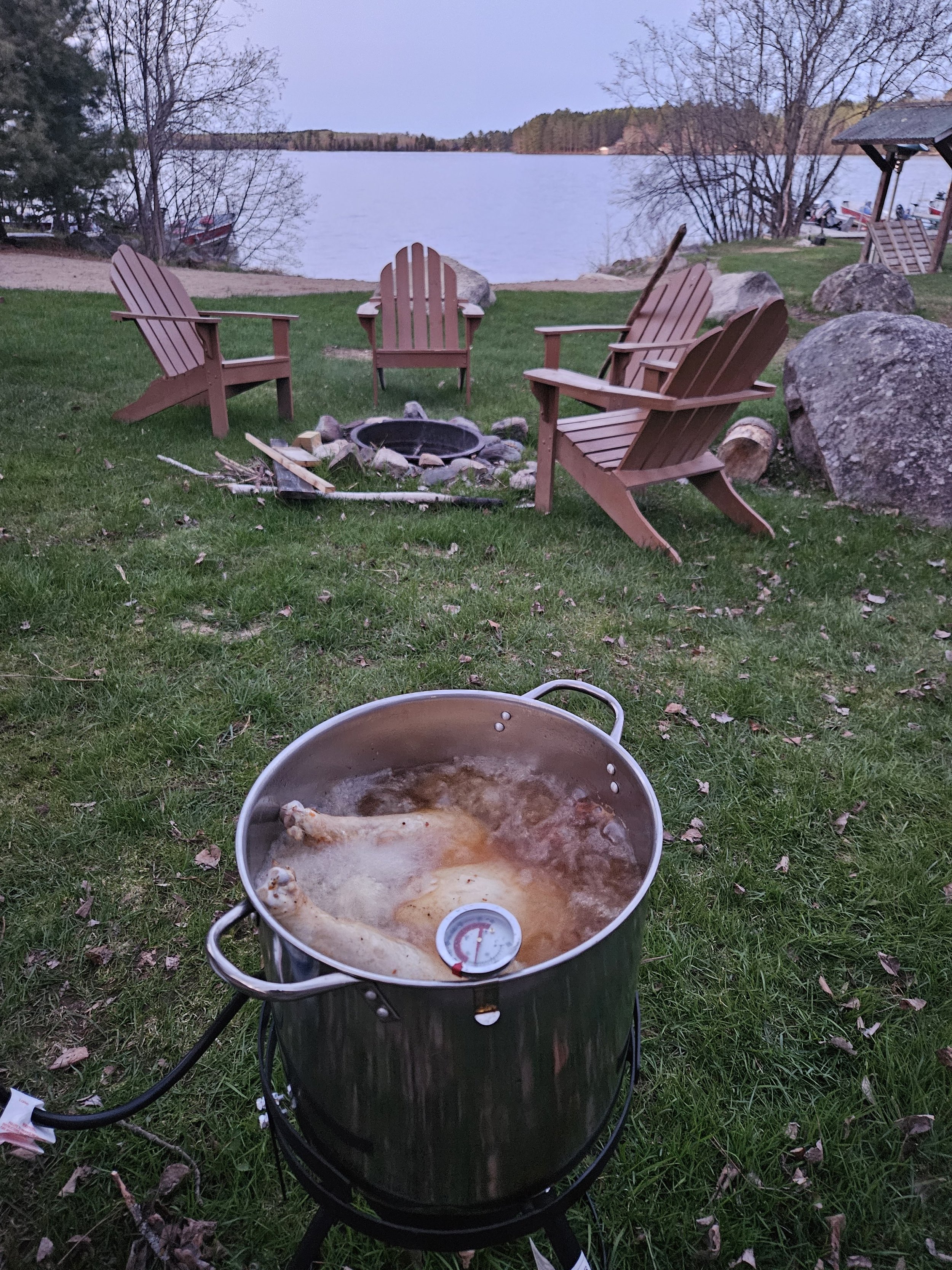 Outdoor scene with a large pot frying a turkey over a gas burner on a grassy lawn, surrounded by Adirondack chairs, a fire pit, and a lake in the background.