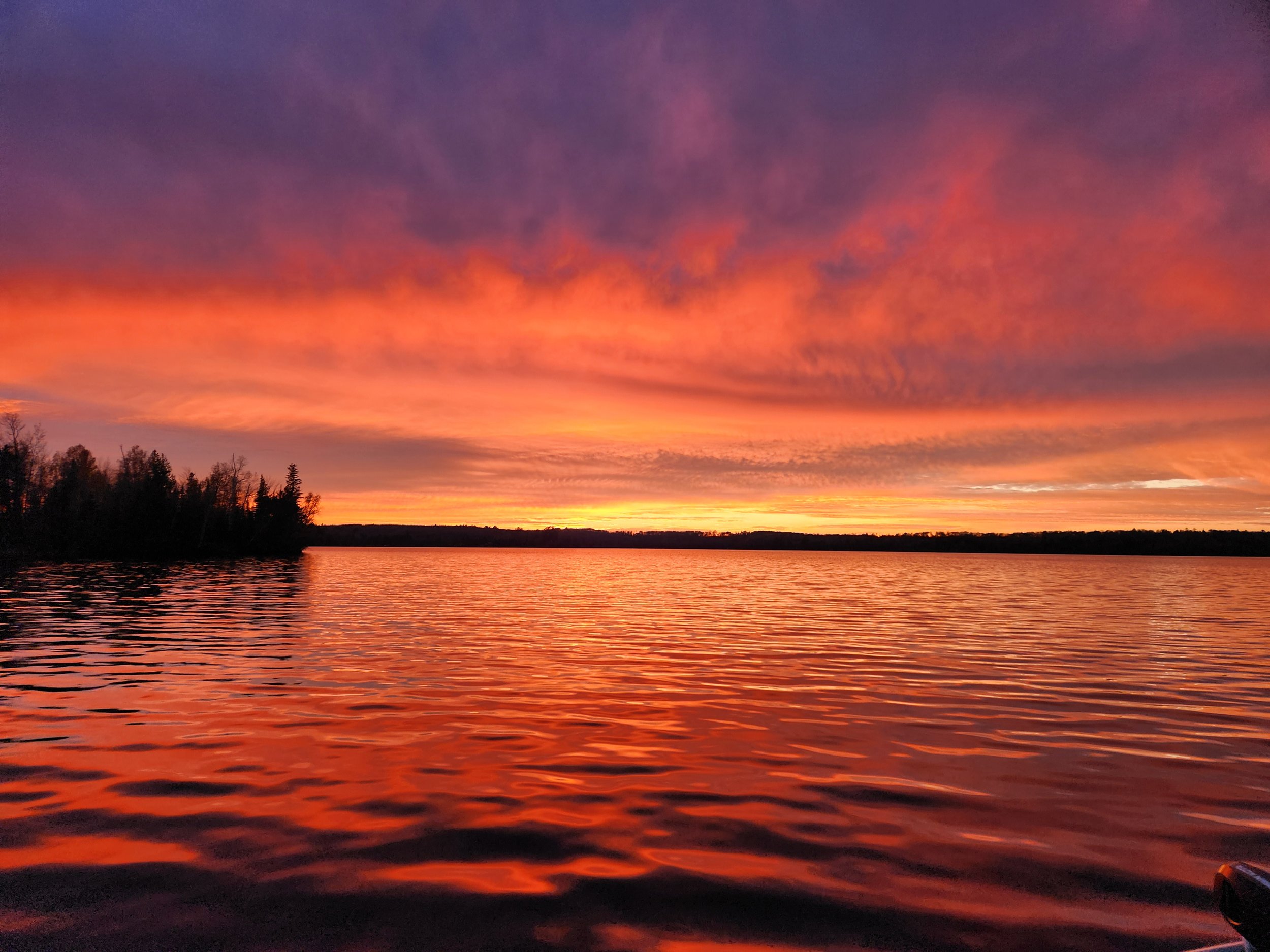 Vibrant sunset over a calm lake with silhouetted trees on the horizon, reflecting orange and pink hues in the water.