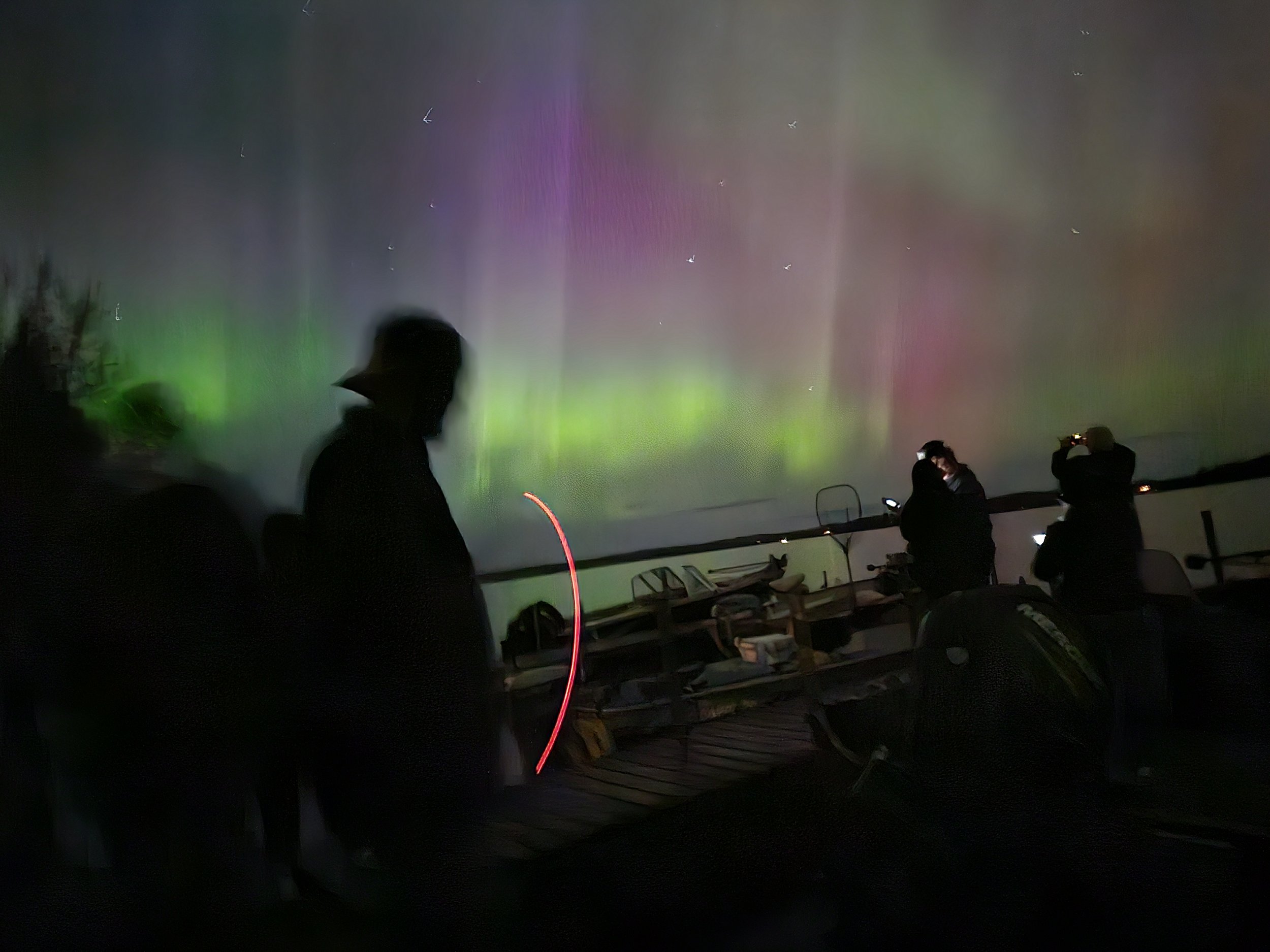 Silhouetted people observing northern lights with green and purple hues over a lake at night, with faint stars visible in the sky.