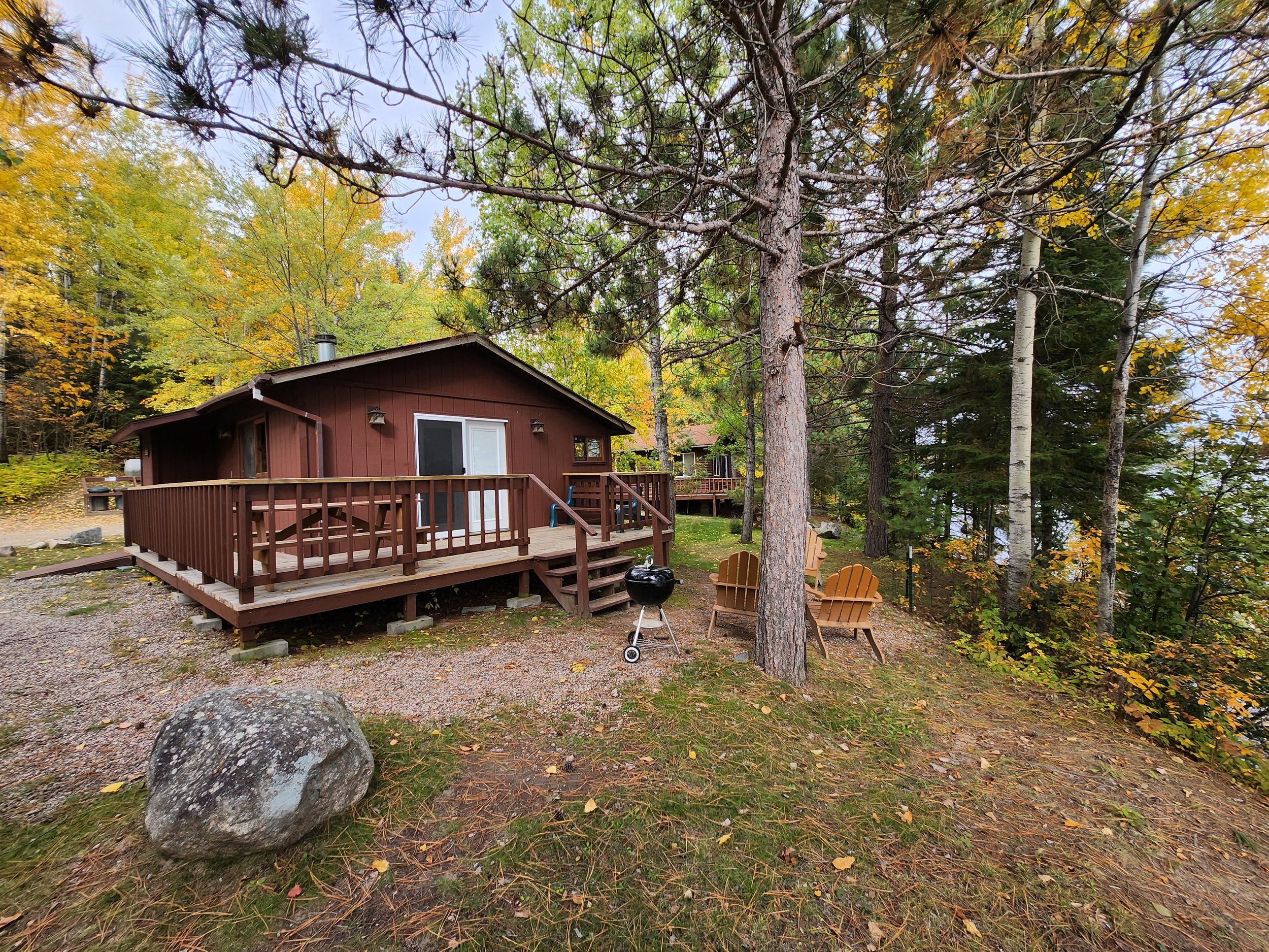 Cabin in wooded area with deck, Adirondack chairs, and a charcoal grill, surrounded by trees with autumn foliage.