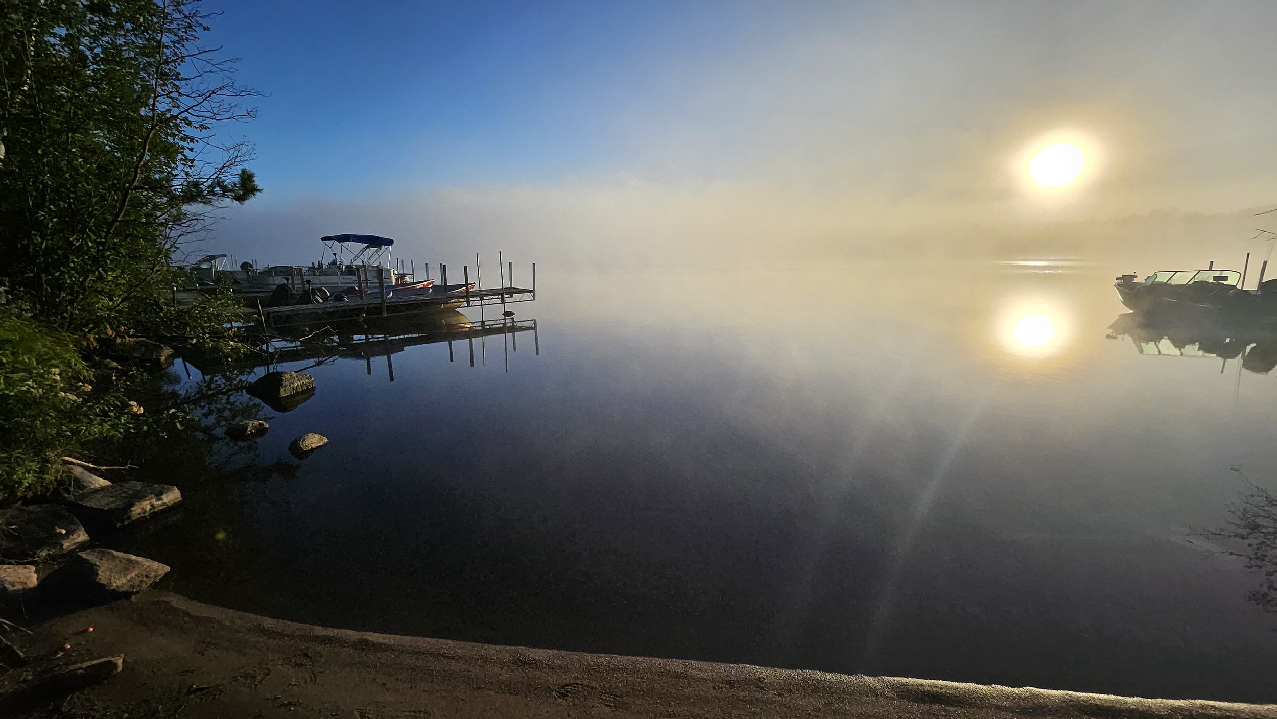 Serene lake at sunrise with fog, boats docked, and a visible sun reflection on the water.