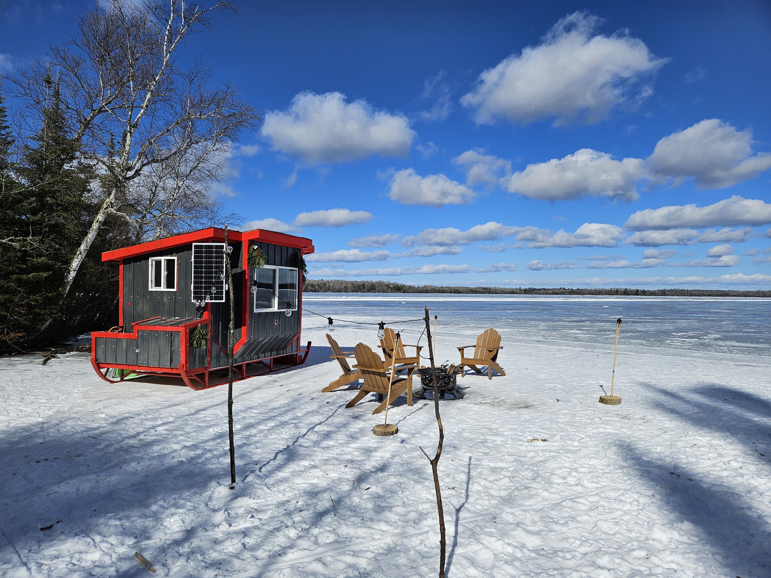 A small cabin with red trim sits on a snowy lakeside, surrounded by trees and under a partly cloudy sky. There are wooden chairs and a fire pit set up on the ice-covered lake.