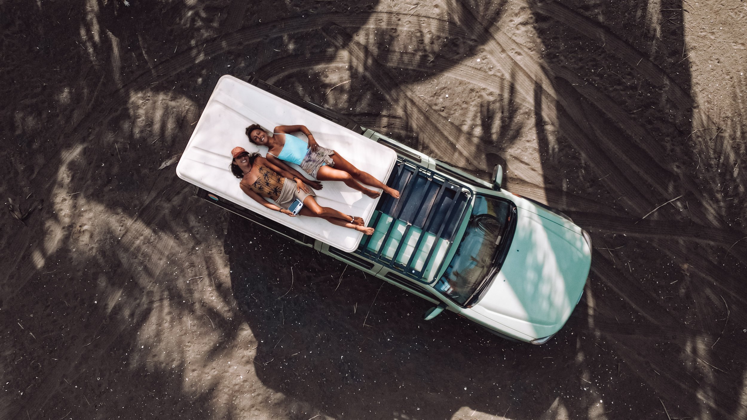 Two people lying on top of a car in a beach setting, with sand and palm tree shadows visible.