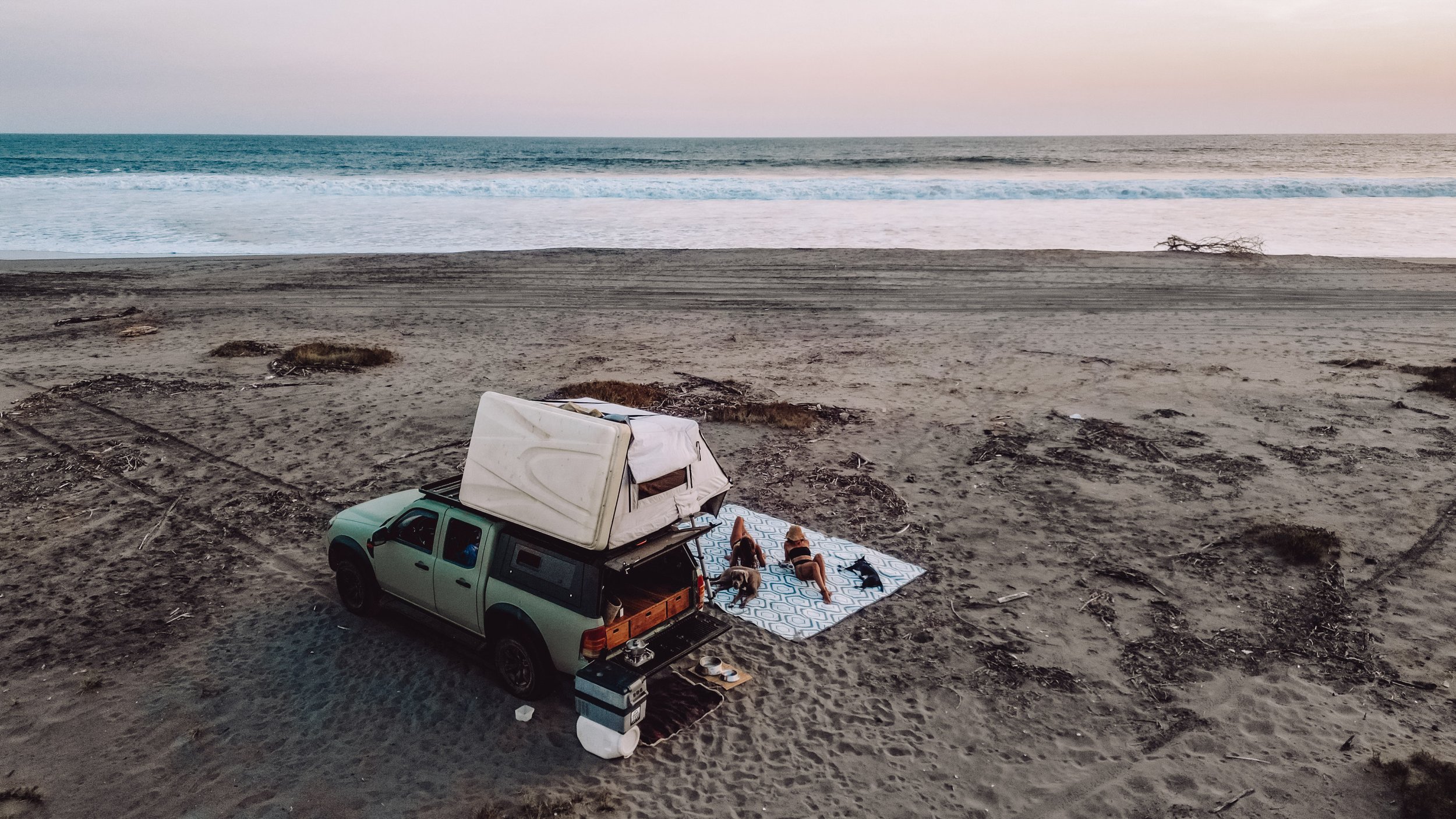 Beach camping scene with a truck and rooftop tent, people relaxing on a blanket, ocean in the background.