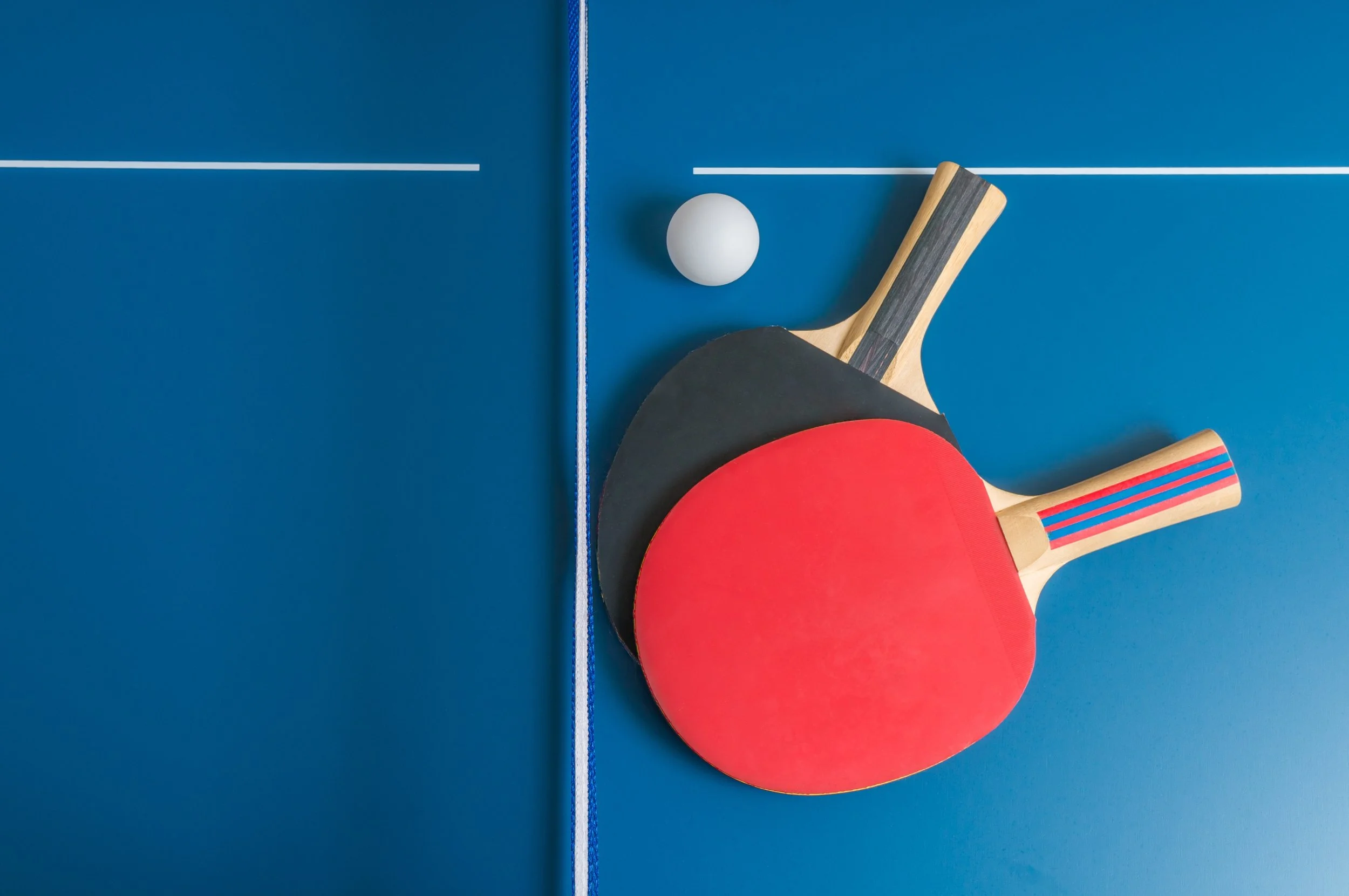 Two ping pong paddles, one with a black surface and one with a red surface, placed on a blue ping pong table with a white ping pong ball nearby.