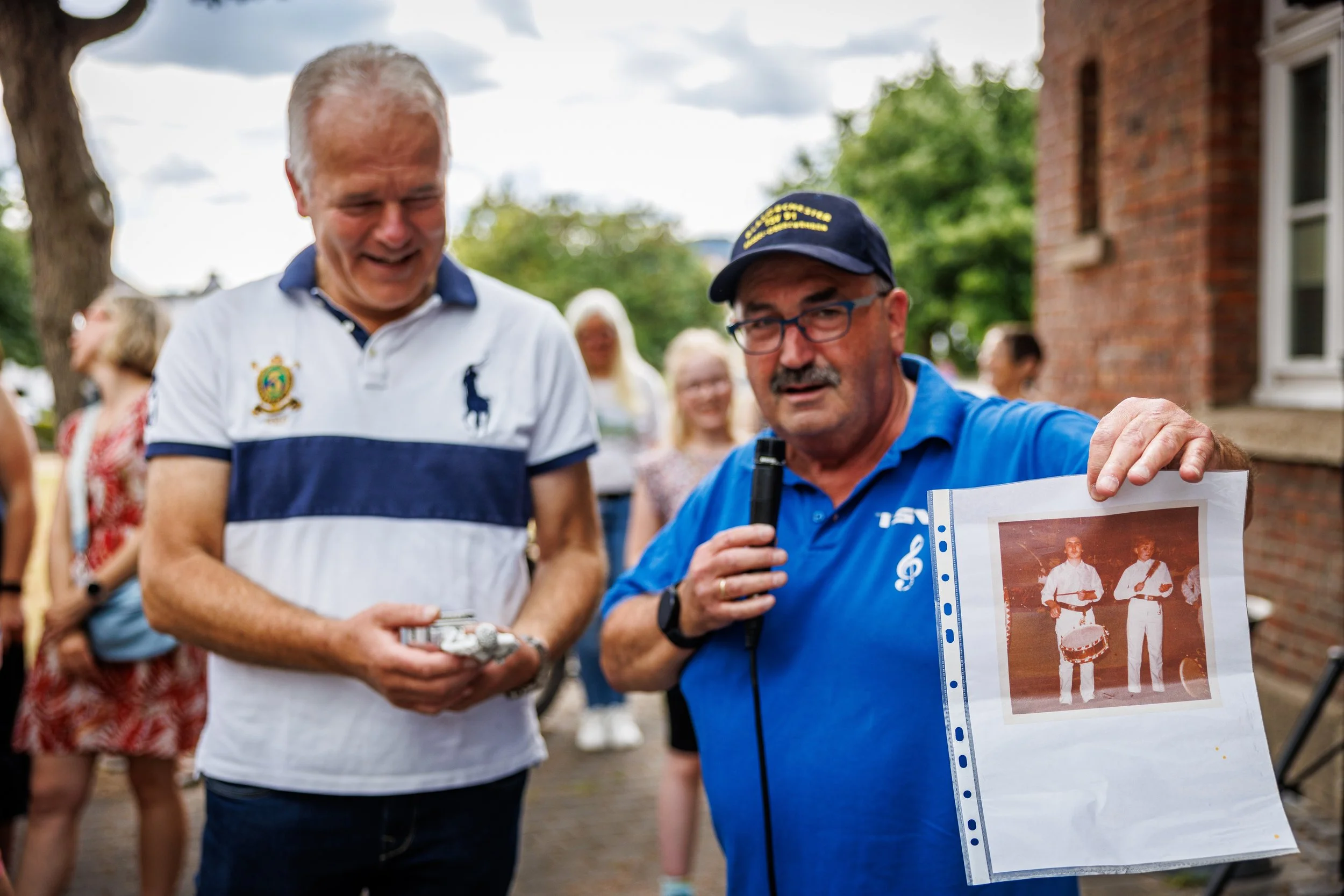 Mann zeigt ein altes Foto von zwei Männern mit Trommel, während eine andere Person neben ihm lächelt, bei einer ausgelassenen Veranstaltung im Freien.