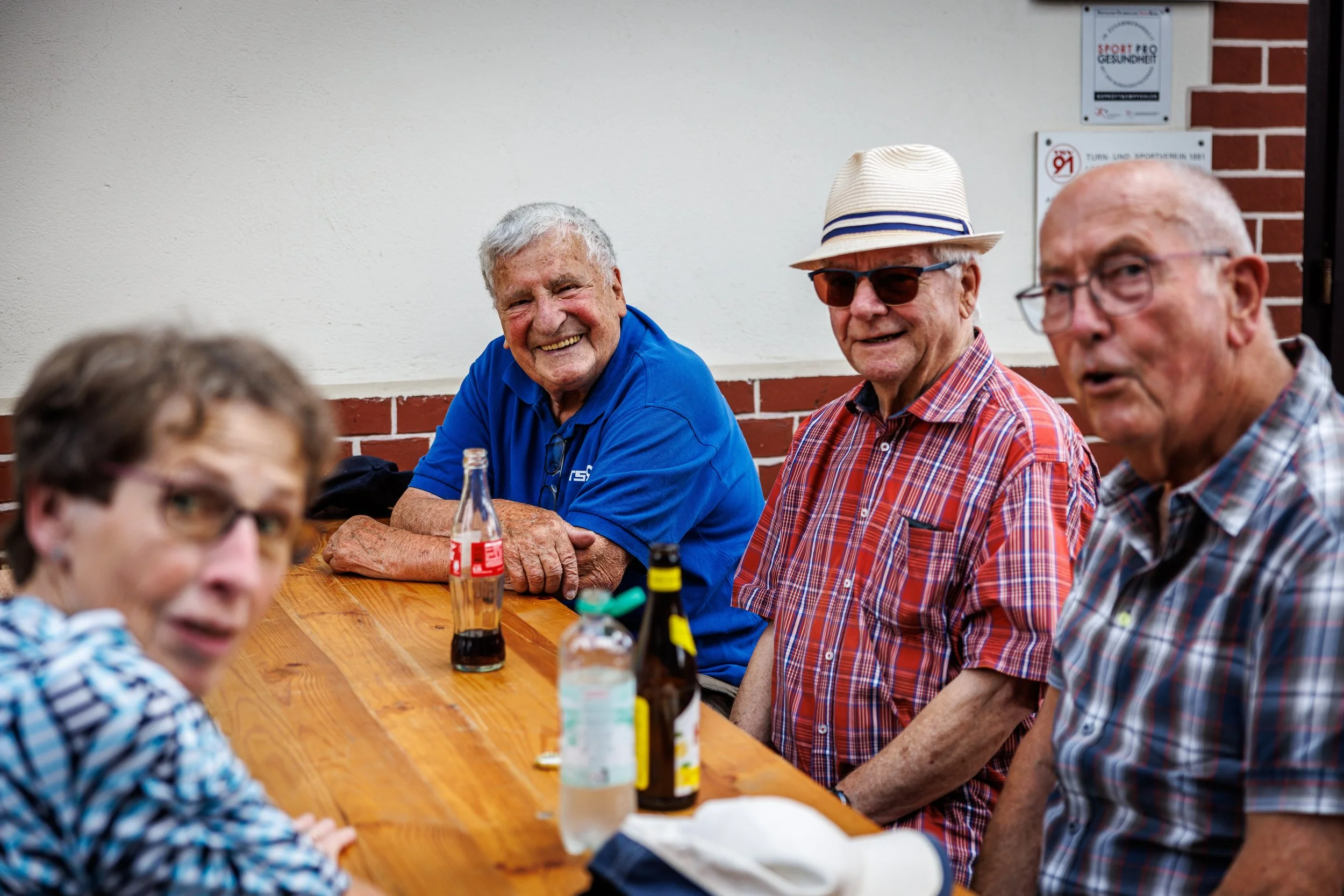 Fünf ältere Menschen sitzen lachend an einem Holztisch bei einem Treffen, Getränke auf dem Tisch, im Hintergrund eine weiße Wand mit Hinweisschildern.