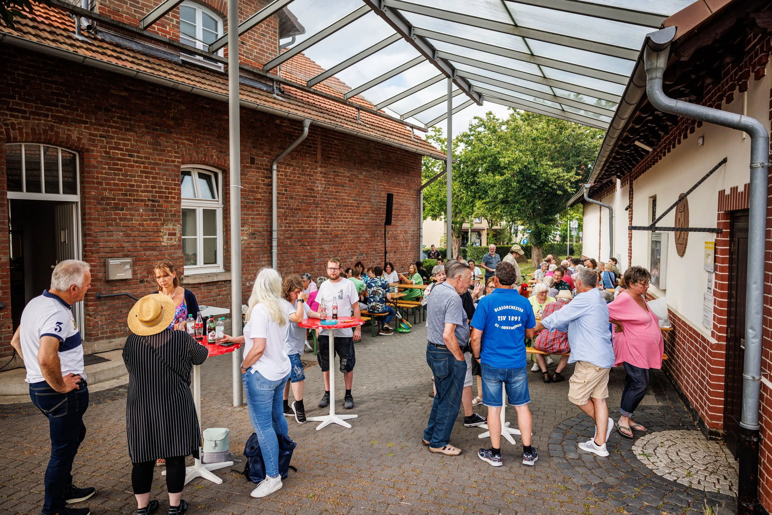 Menschen versammeln sich bei einem Außenevent, einige stehen an Tischen, andere sitzen auf Bänken in einem Innenhof zwischen alten roten Backsteingebäuden mit begrüntem Hintergrund.