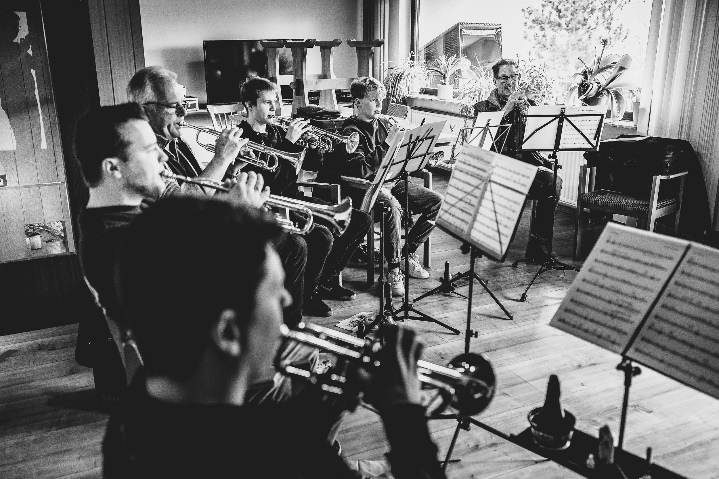 Eine Gruppe von jungen und älteren Musikern, die eine Blaskapelle bildet, spielt gemeinsam in einem Raum mit Holzboden und großen Fenstern. Sie spielen Blasinstrumente, darunter Trompeten. Es gibt Notenständer mit Noten, und im Hintergrund befinden s
