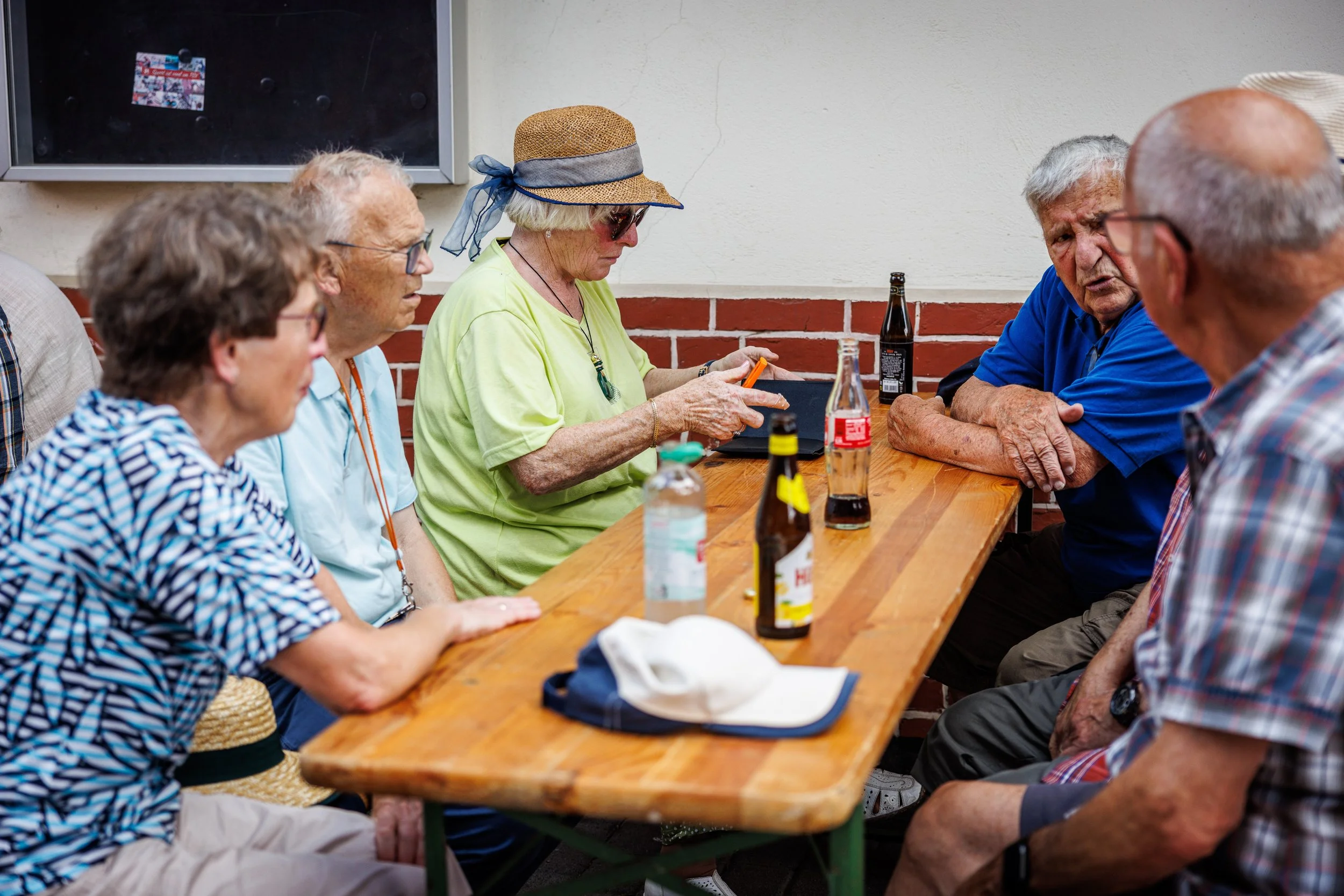 Gruppe älterer Menschen sitzt an einem Holztisch, unterhalten sich und trinken Getränke, im Hintergrund eine weiße Wand.