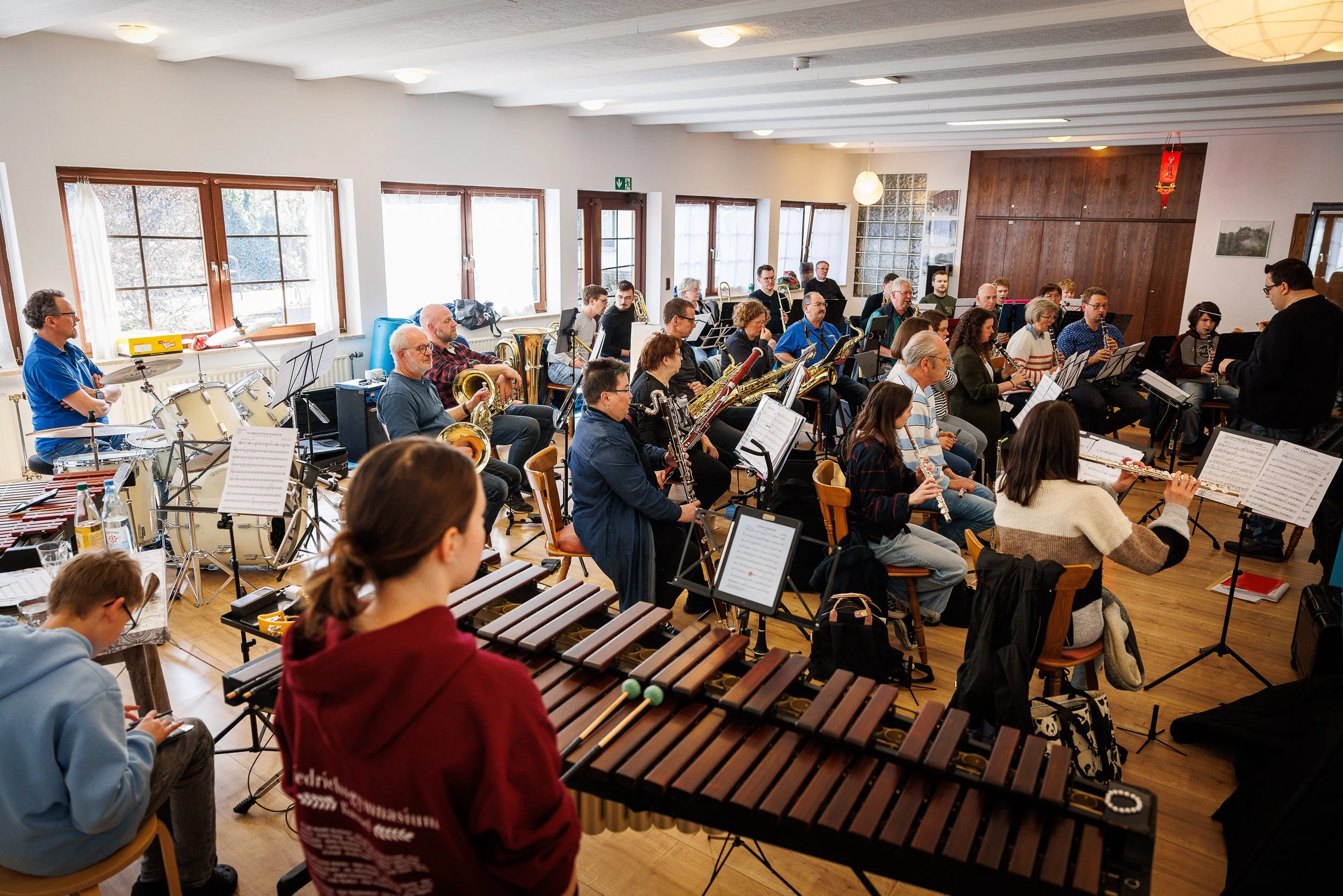 Ein Orchester probt in einem gut beleuchteten Raum mit mehreren Fenstern, Holzfußboden und Off-White-Wänden, während der Dirigent den Takt vorgibt.
