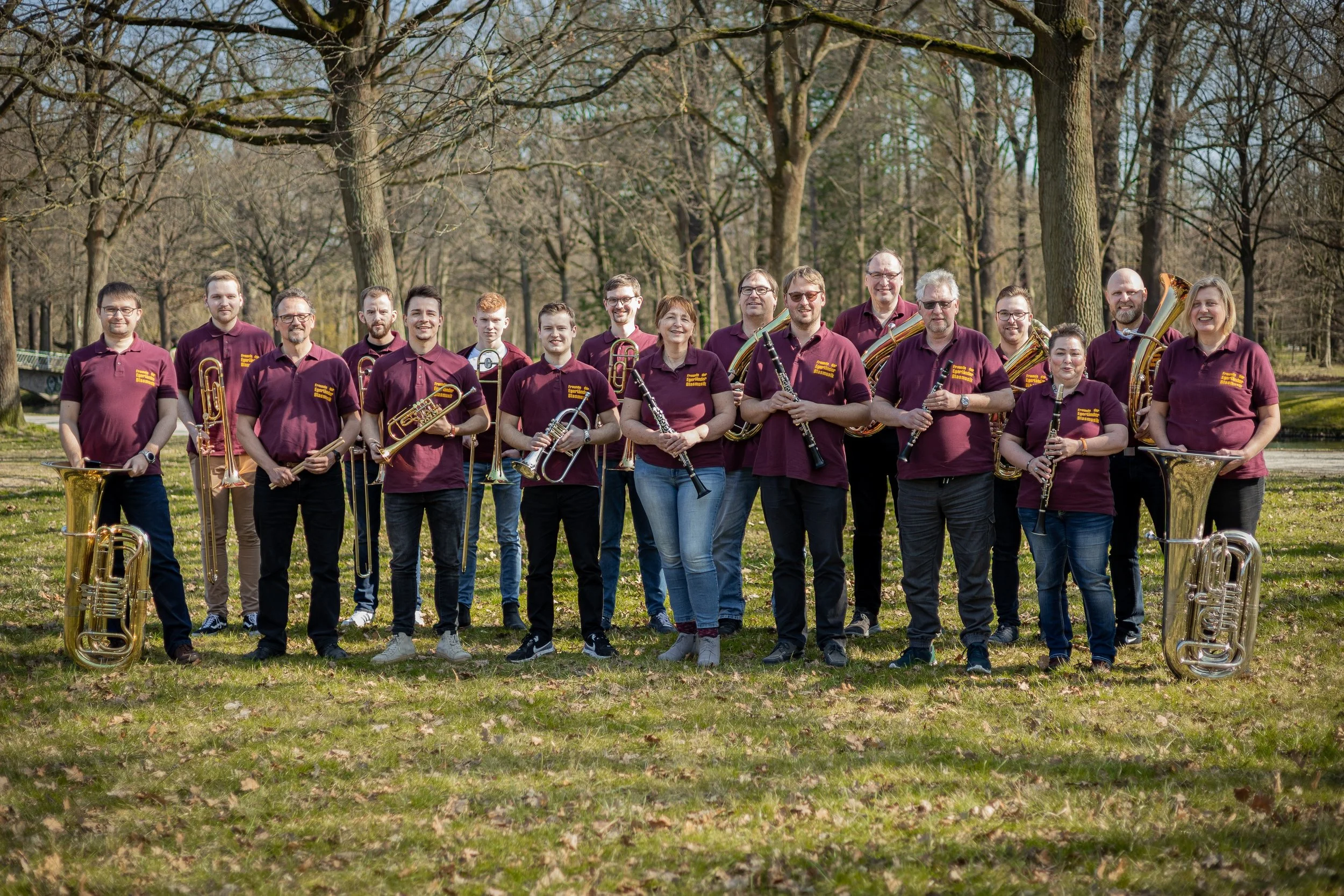 Eine Gruppe von 15 Musikern steht im Freien auf einer Wiese, alle tragen burgunderfarbene Hemden mit gelbem Text, einige halten Blasinstrumente wie Trompeten, Klarinetten und Fagotte.