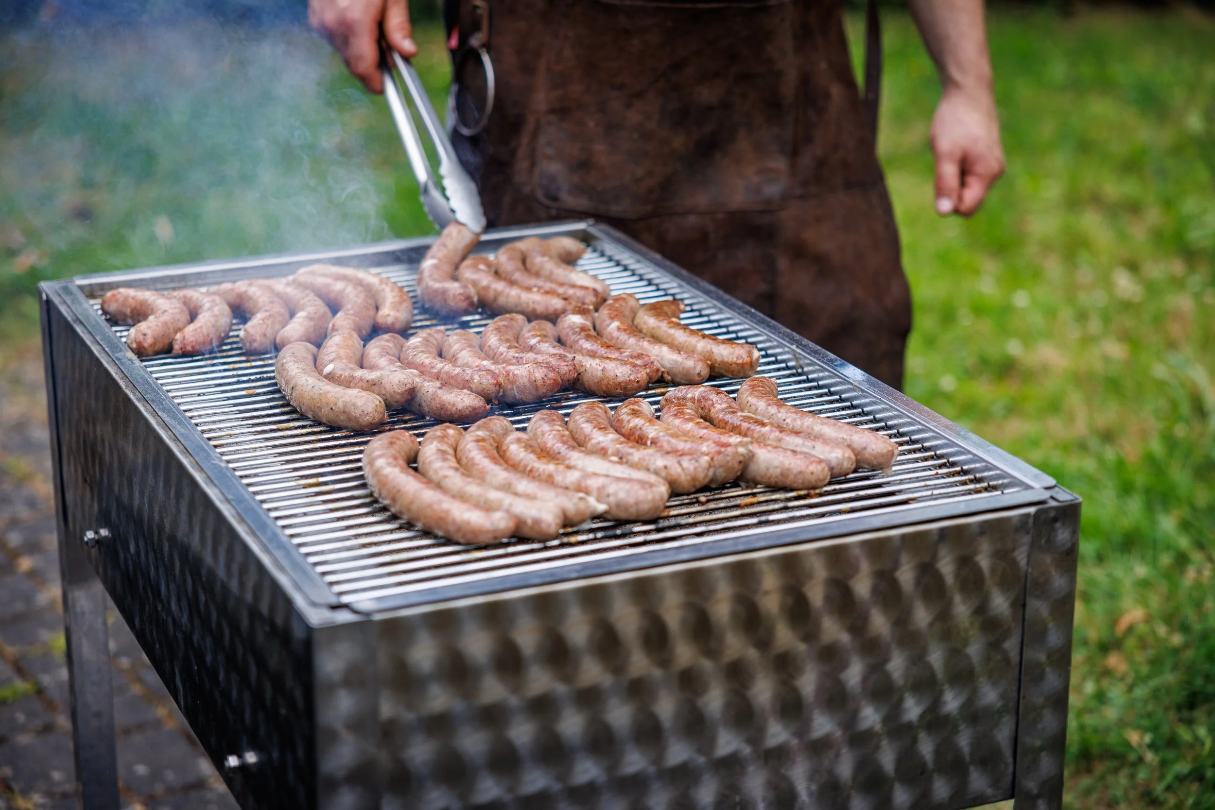 Grill mit mehreren Bratwürsten, jemand benutzt Zange, um die Würste zu wenden, im Freien
