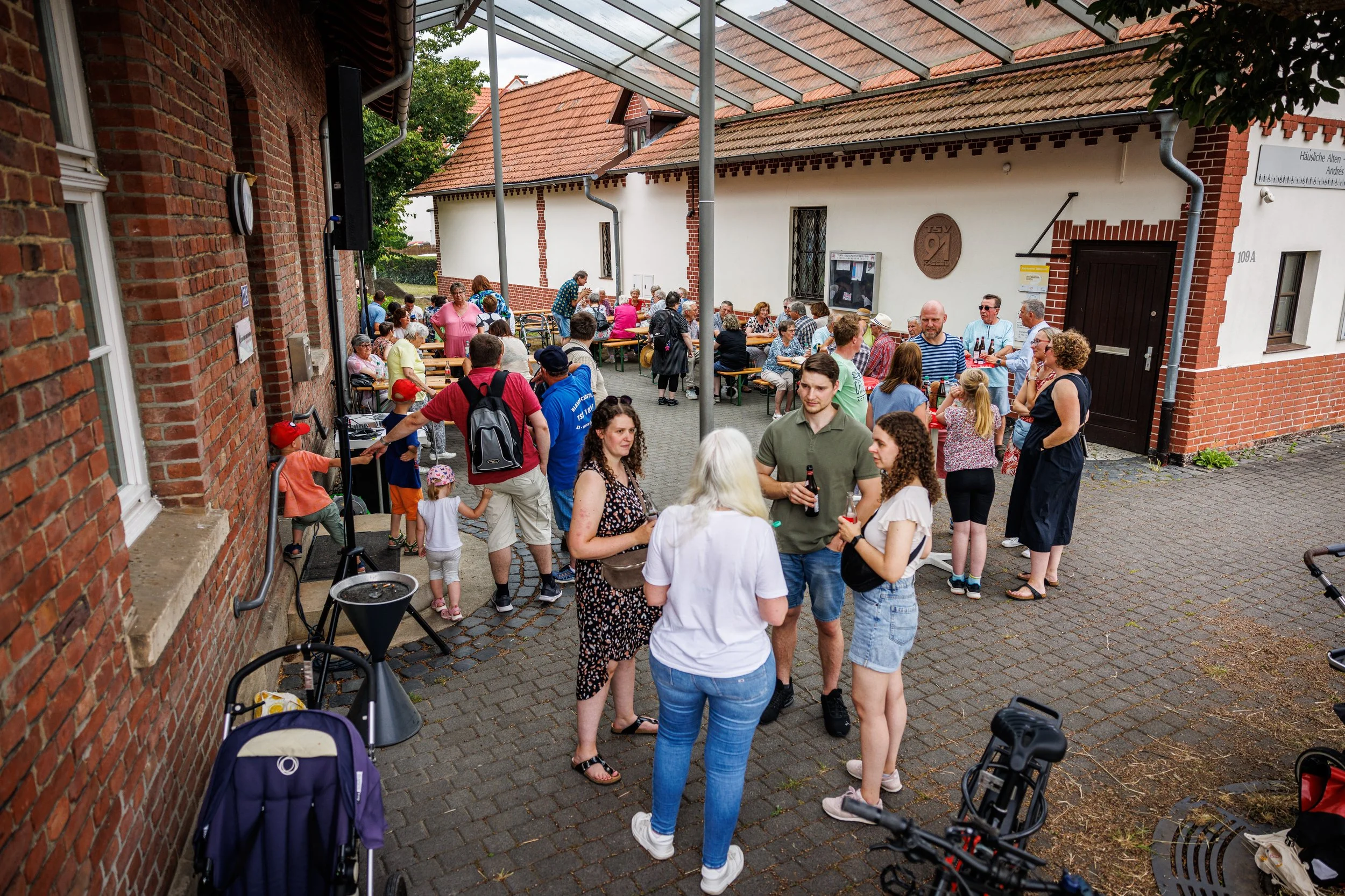 Menschen versammeln sich auf einem Hof im Freien, einige sitzen an Tischen, andere stehen und unterhalten sich. Es scheint eine soziale Veranstaltung zu sein, mit etwa 30 Personen, darunter Kinder.
