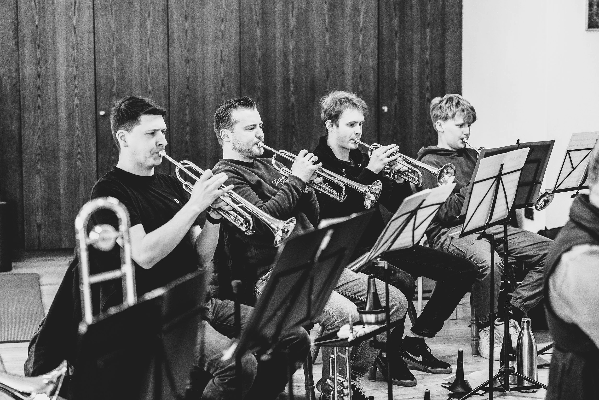Fünf junge Männer spielen in einem Orchester Geige und andere Blasinstrumente, sitzen in einem Raum mit Holzwand im Hintergrund.