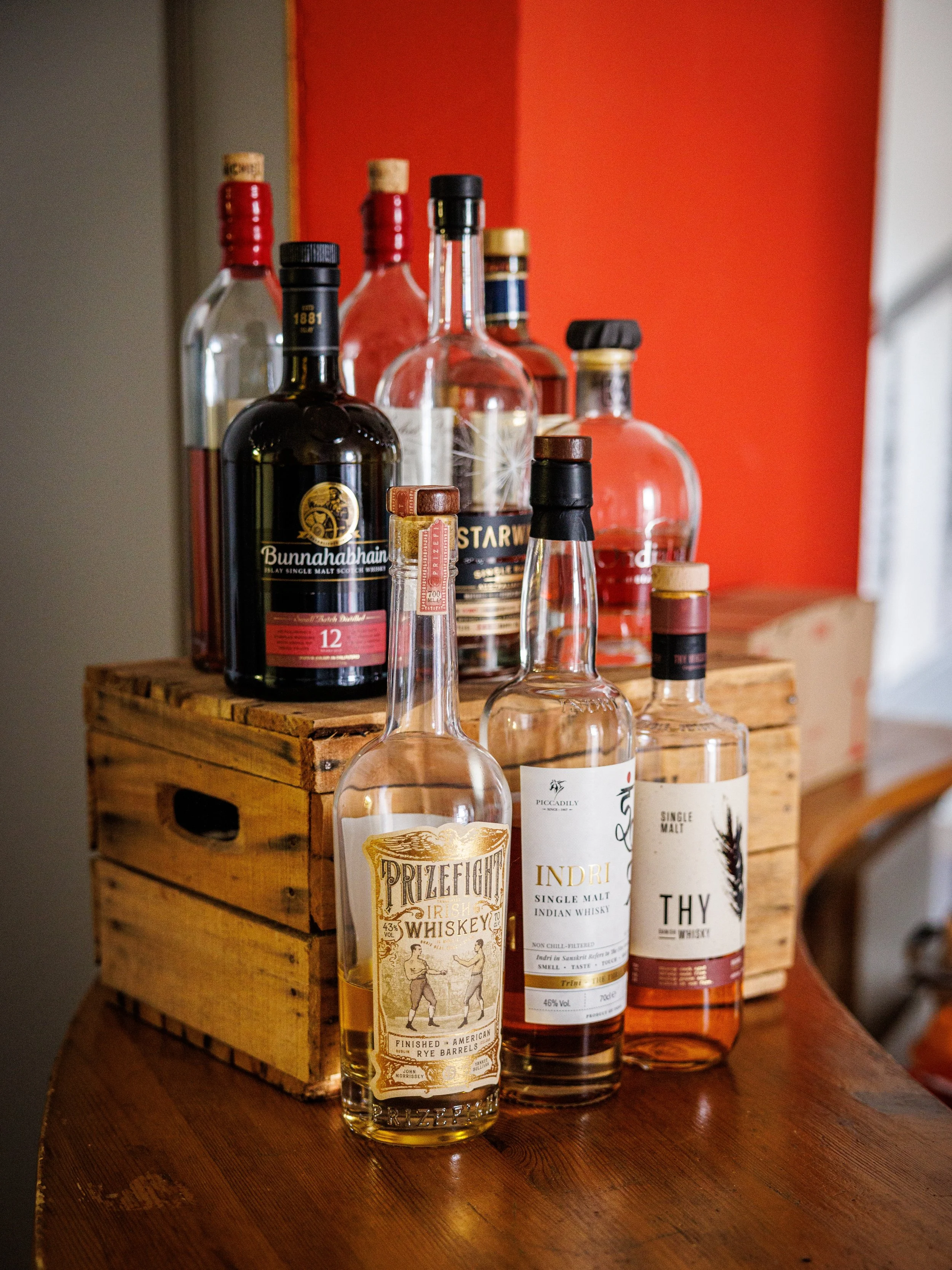 Verschiedene Flaschen mit alkoholischen Getränken, darunter Whisky und andere Spirituosen, stehen auf einem Holztisch und in einer Holzkiste, vor einer roten Wand.