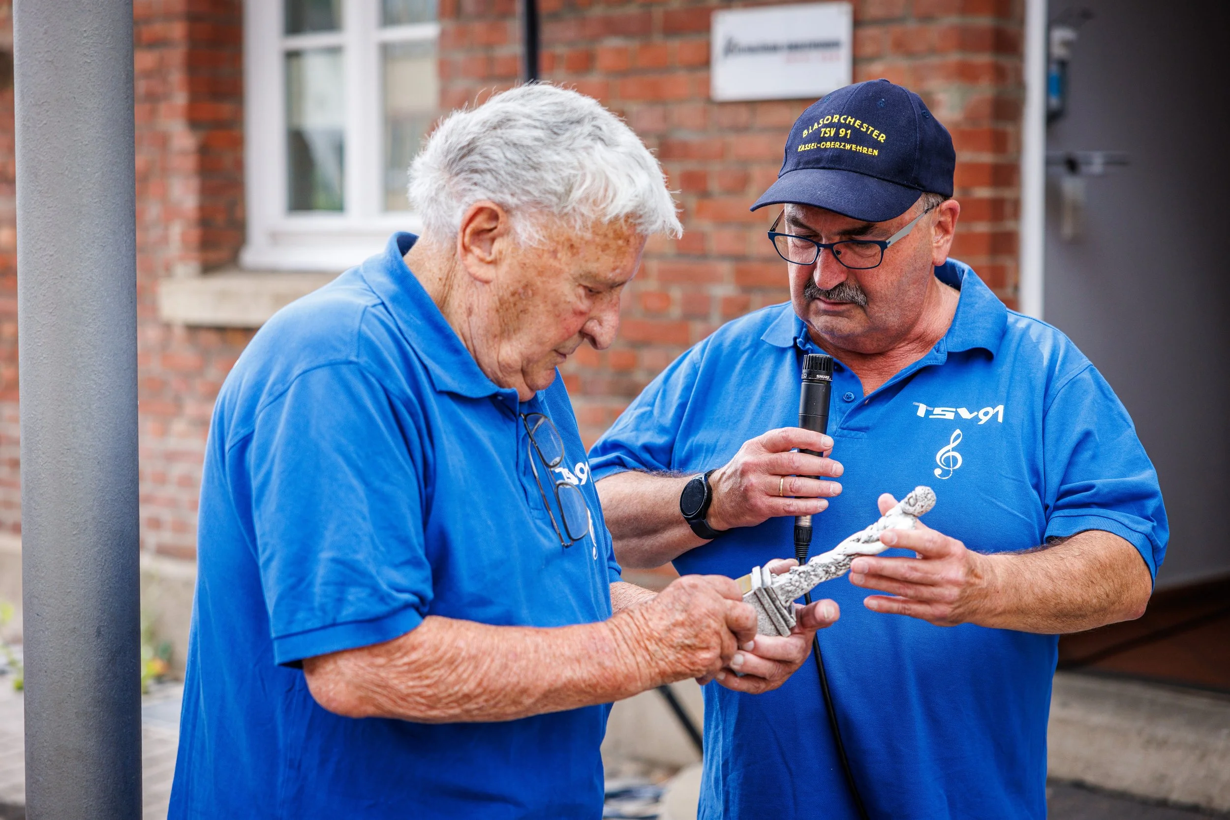 Zwei Männer in blauen T-Shirts mit TS91-Aufdruck betrachten eine Skulptur, während einer ein Mikrofon hält.