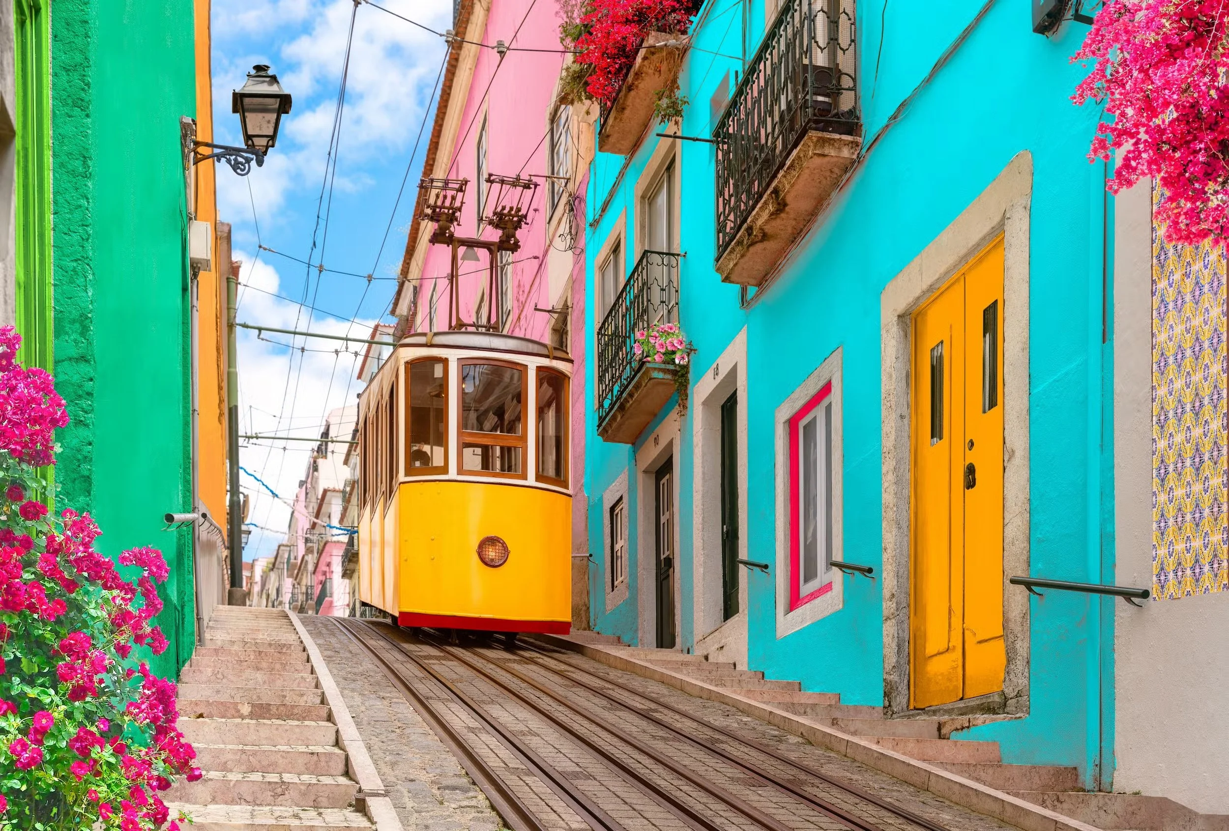 Colorful street with pink, blue, and green buildings, flower pots on balconies, a yellow tram on a steep track, and pink flowers on the left.