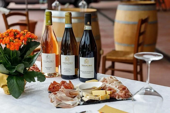 A table set with a flower arrangement, three bottles of wine, and a cheese and meat platter, with empty glasses and a wine barrel in the background.