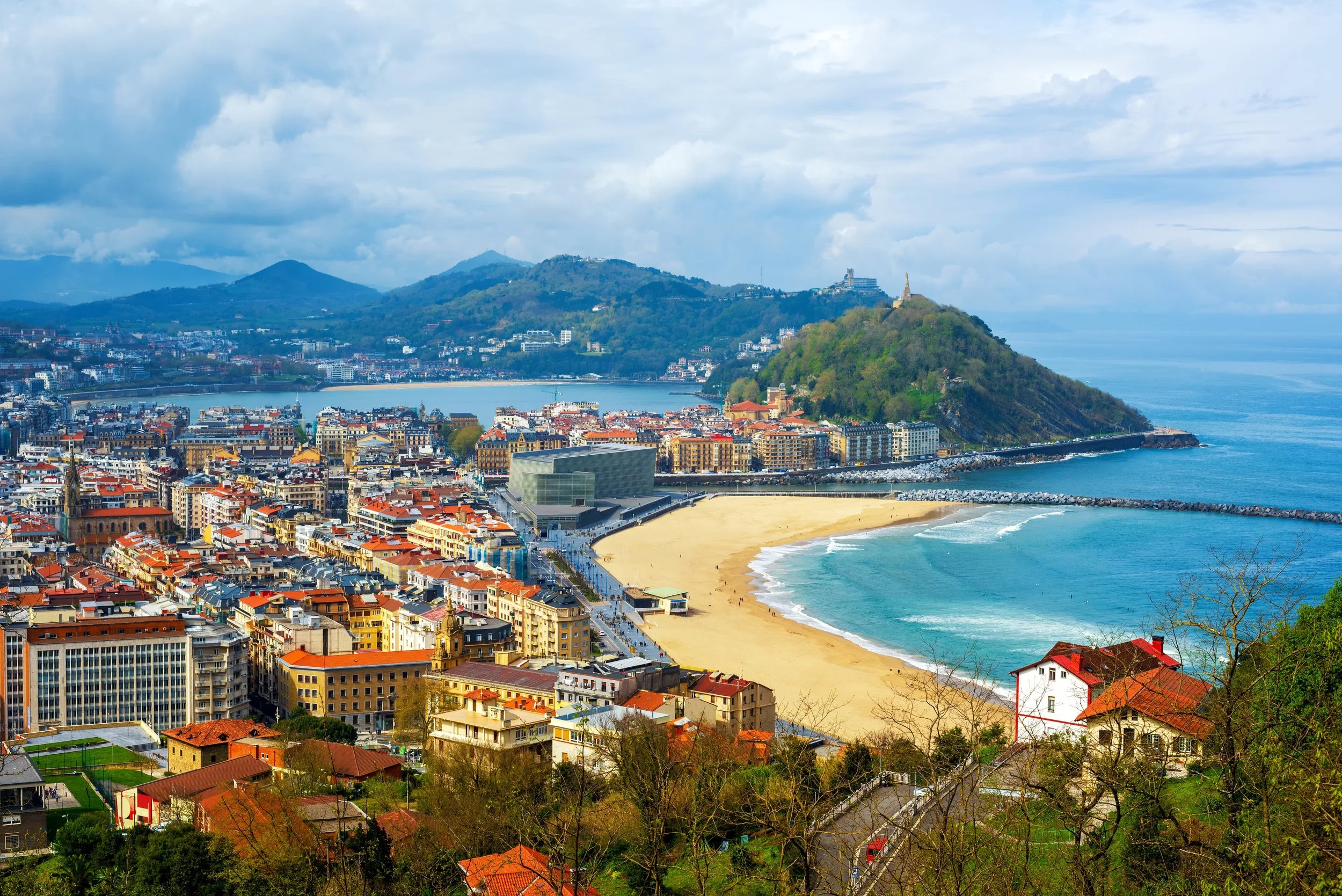Panoramic view of a city with a beach, mountains, and water. Colorful buildings line the shore, with a sandy beach and a breakwater extending into the water. Hills and green areas are in the background under a partly cloudy sky.