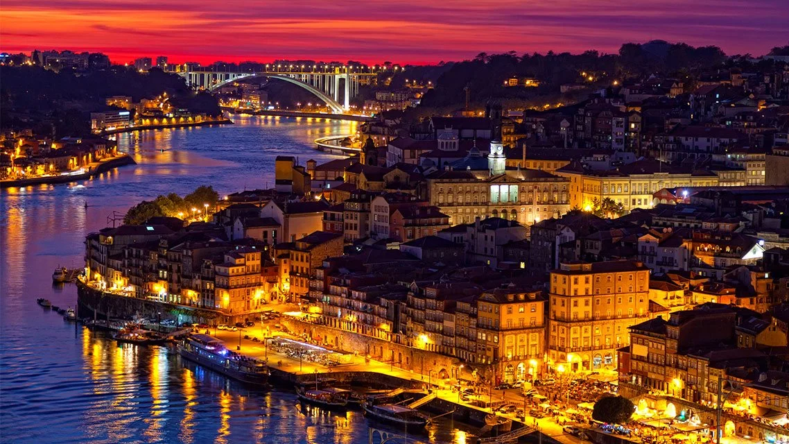 Cityscape of a European city at dusk, illuminated buildings along a river with a bridge and colorful sunset sky.