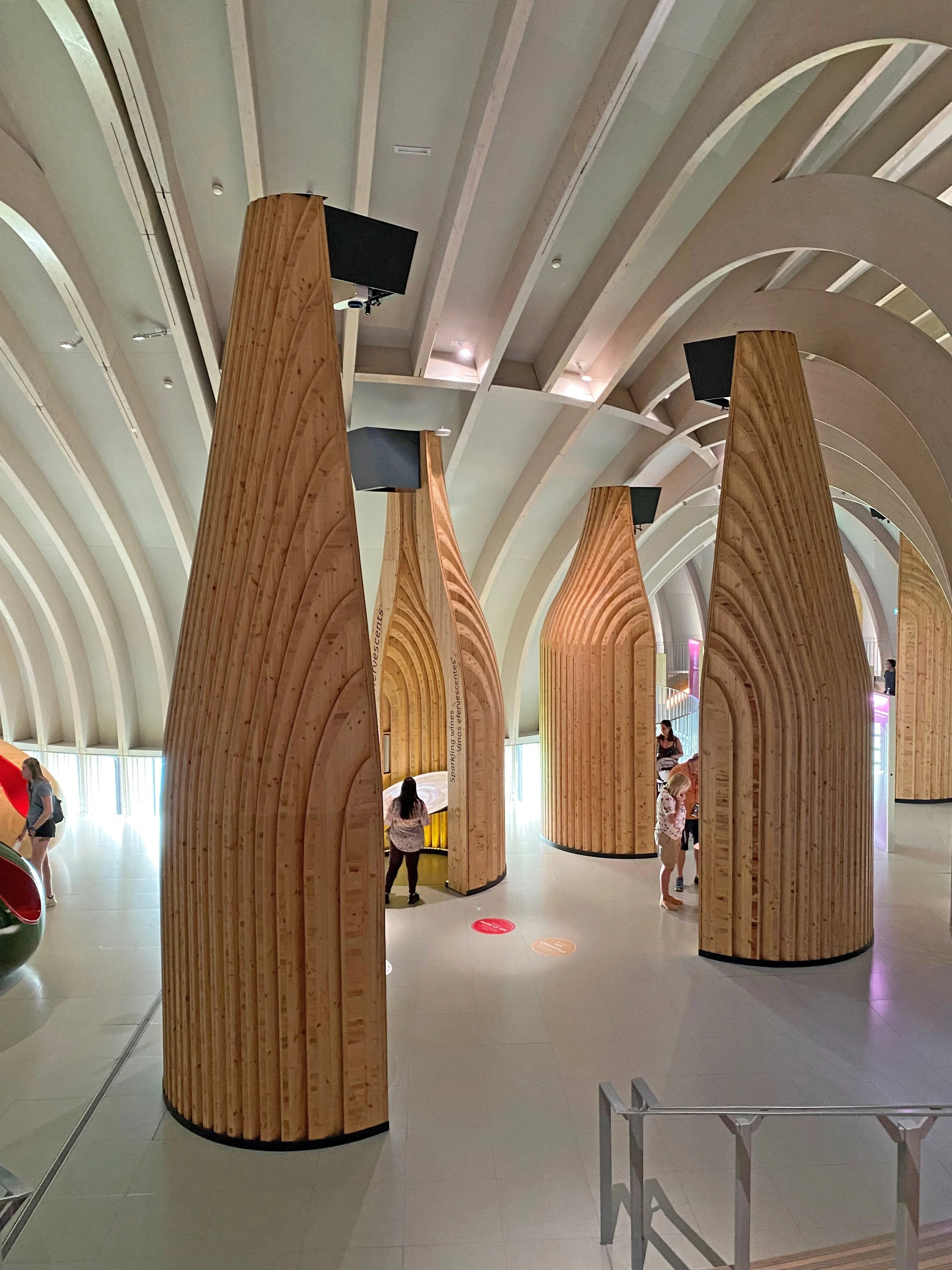 Interior view of a modern building with large, curved wooden structures and a high arched ceiling, featuring people walking and standing.