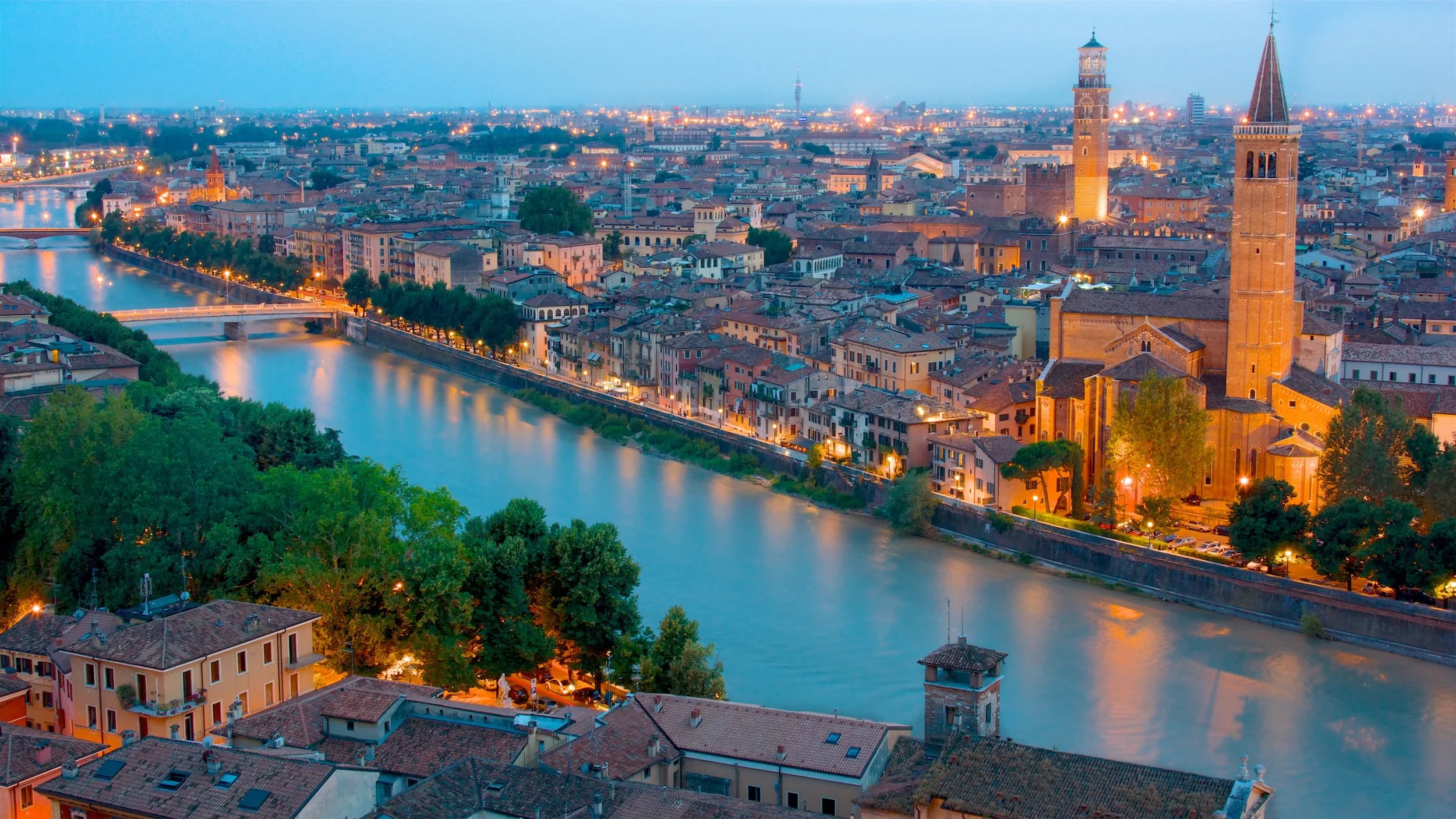 Aerial view of a city with a river running through it, illuminated buildings at dusk, and prominent church towers.