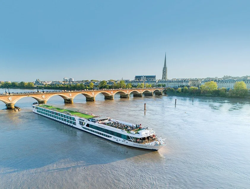 A large river cruise boat sailing on the river with a cityscape and a bridge in the background under a clear blue sky.