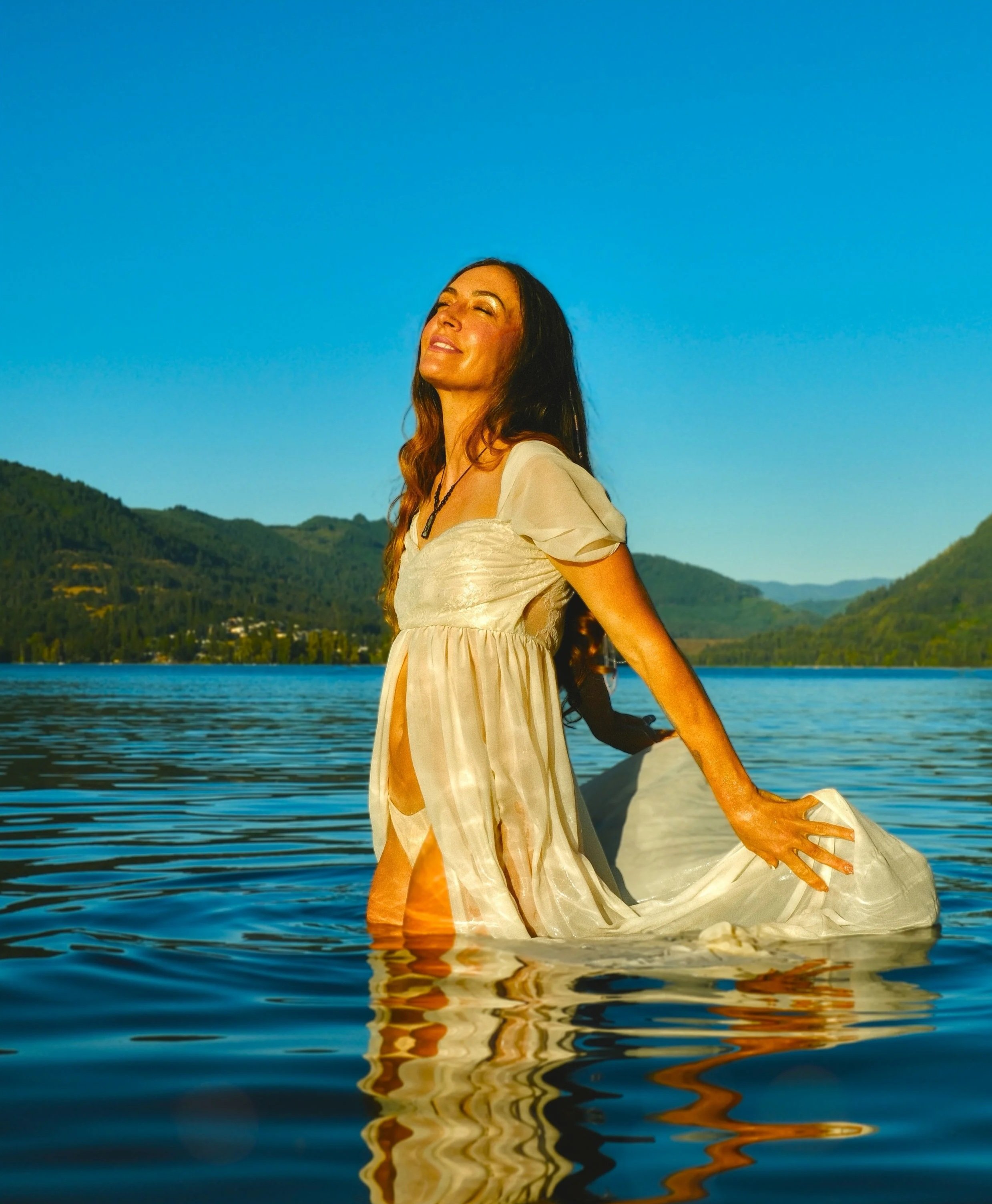 A woman with long dark hair standing in a body of water near a mountain range, with a clear blue sky above, wearing a light-colored flowing dress and smiling with her eyes closed.
