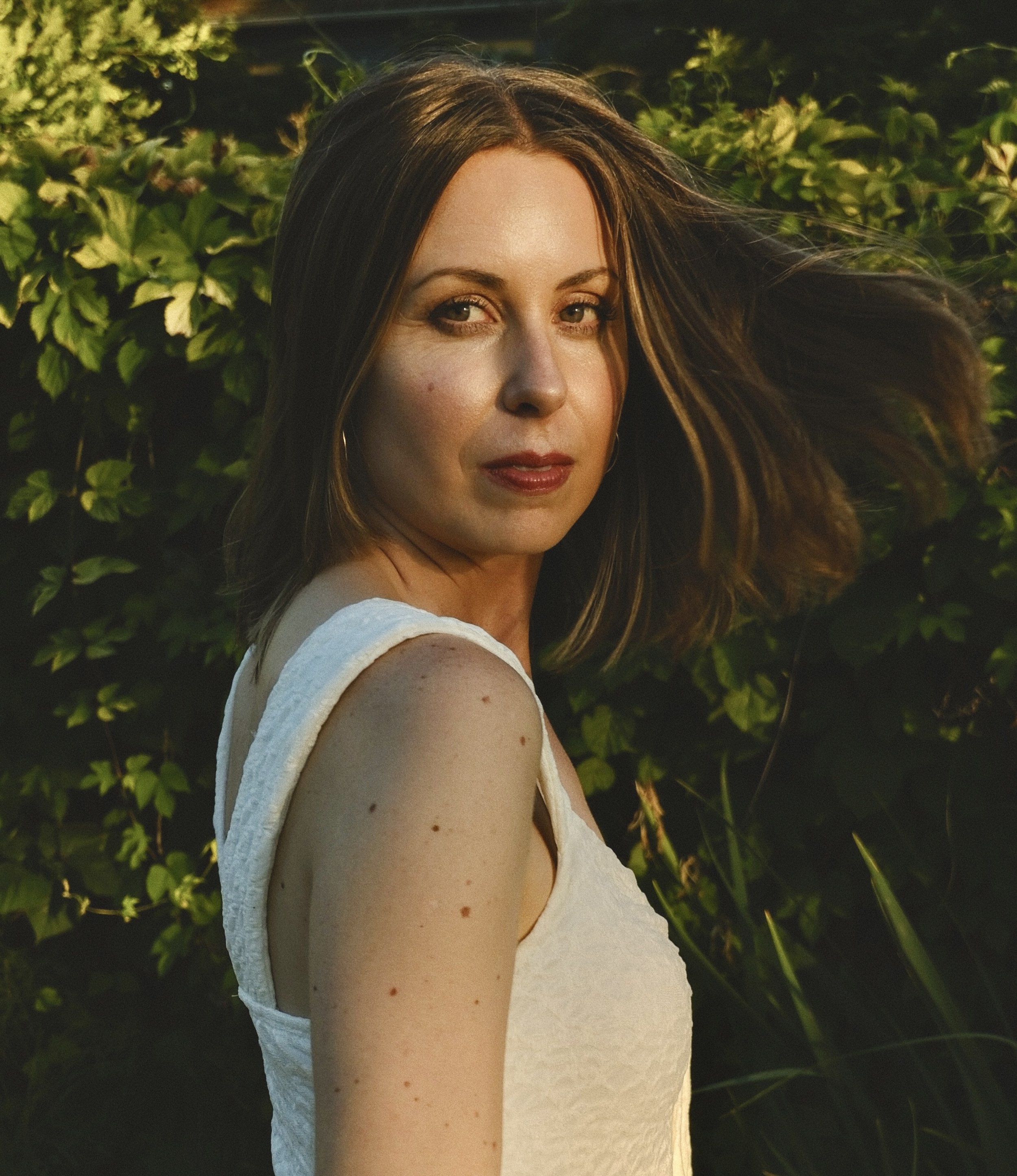 A woman with brown hair and light skin looking over her shoulder outdoors, with green foliage in the background.
