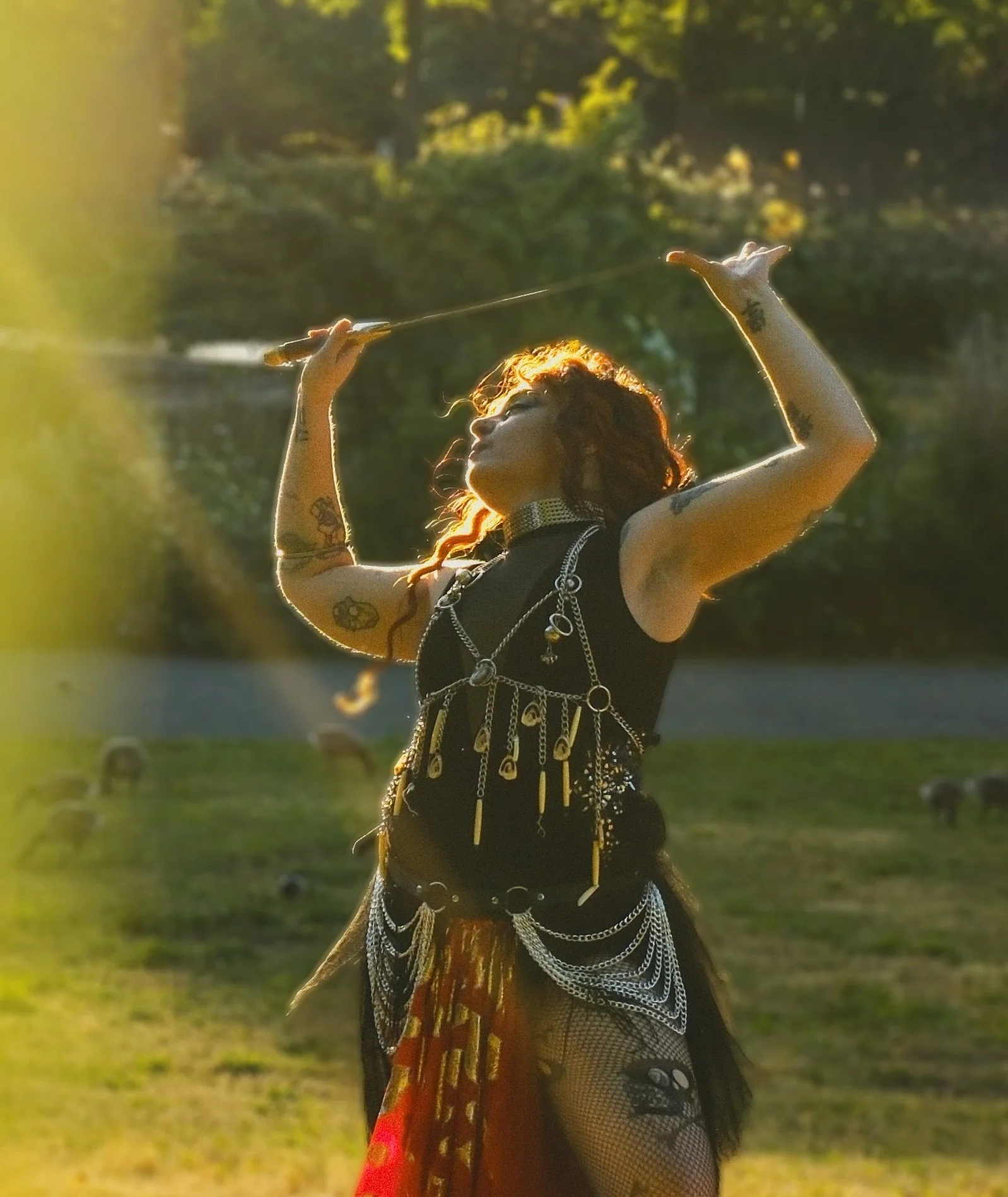 A woman with long, curly hair and tattoos on her arms dressed in black and adorned with silver jewelry and chains, holding a stick and extending her arms in an outdoor setting with trees and a river in the background, illuminated by warm sunlight.