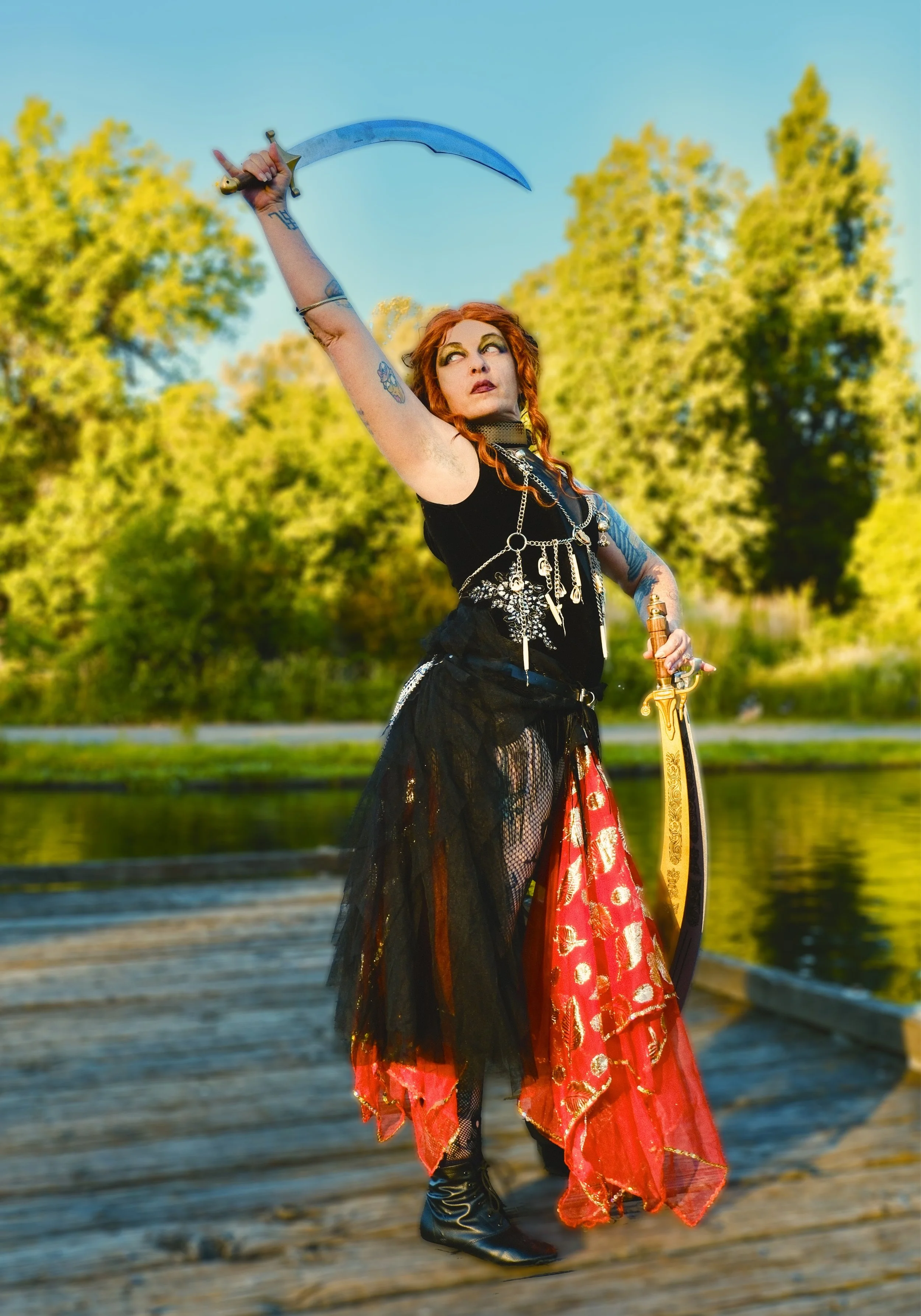 A woman dressed as a pirate with curly red hair holding a sword aloft on a wooden pier with trees and a river in the background.