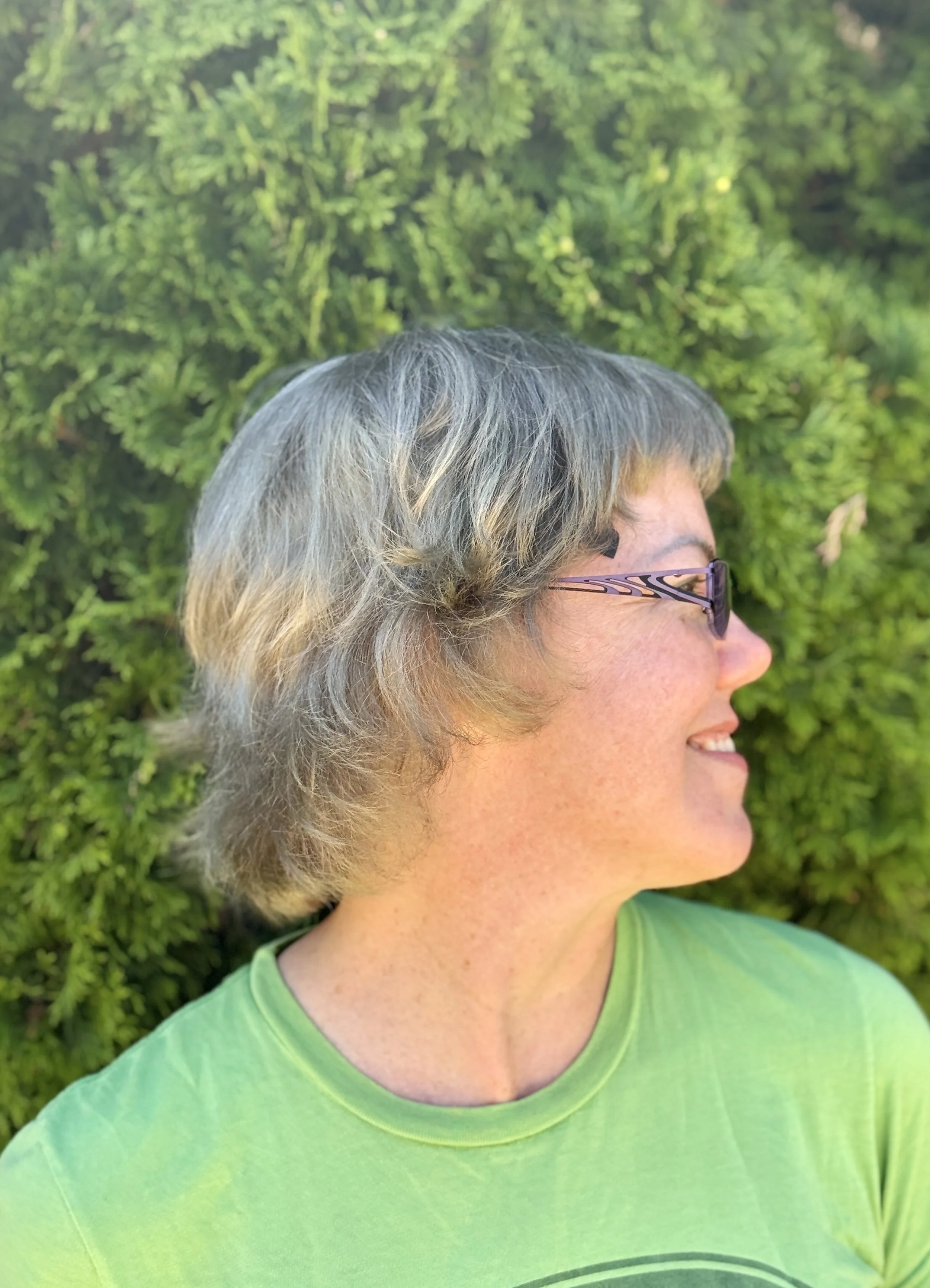 Profile of a woman with shoulder-length gray hair wearing glasses and a green T-shirt, outdoors with a background of green shrubs.