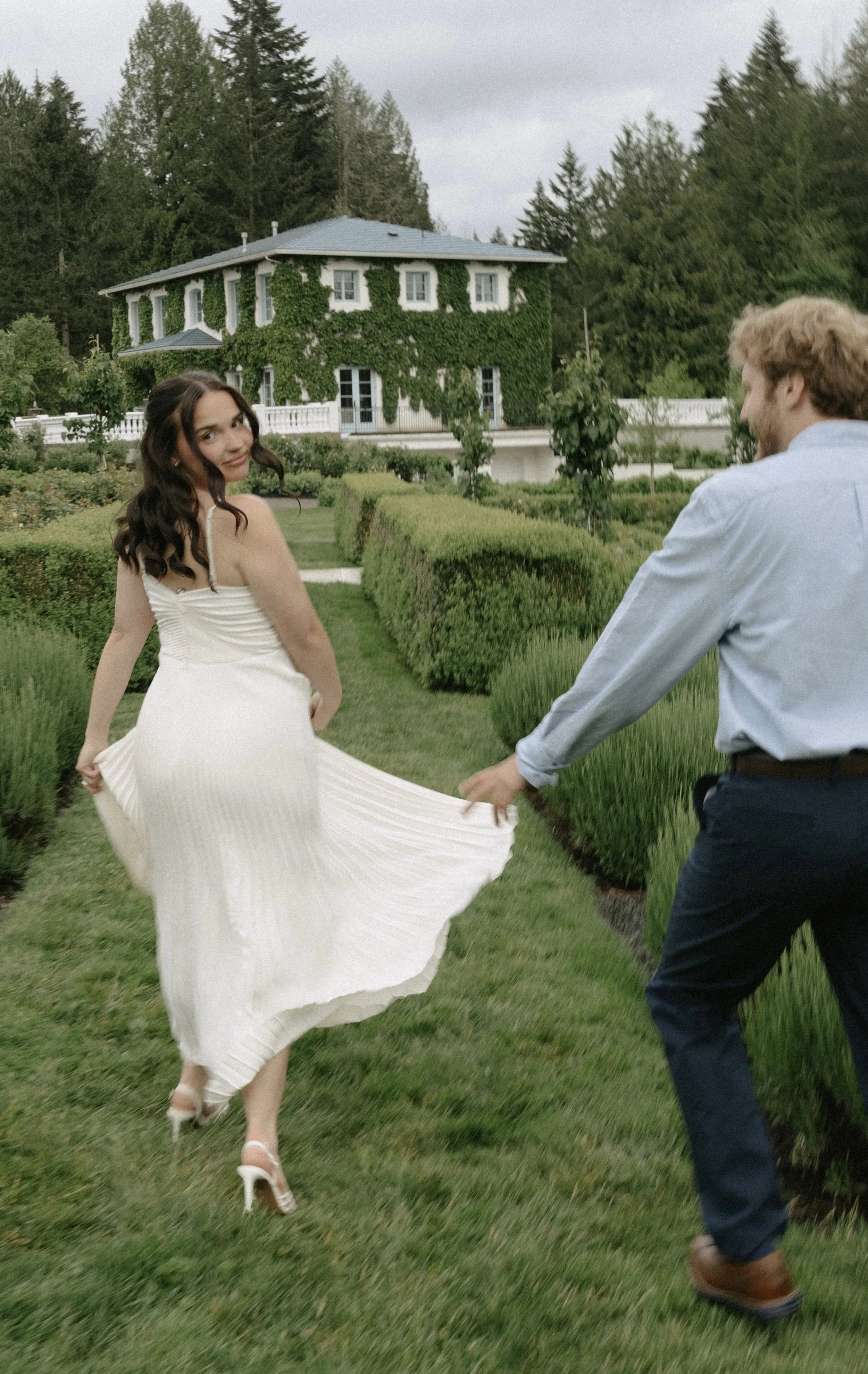 A woman in a white dress and heels dancing in a garden, holding the hand of a man in a light blue shirt and dark pants. A large green ivy-covered house is visible in the background.