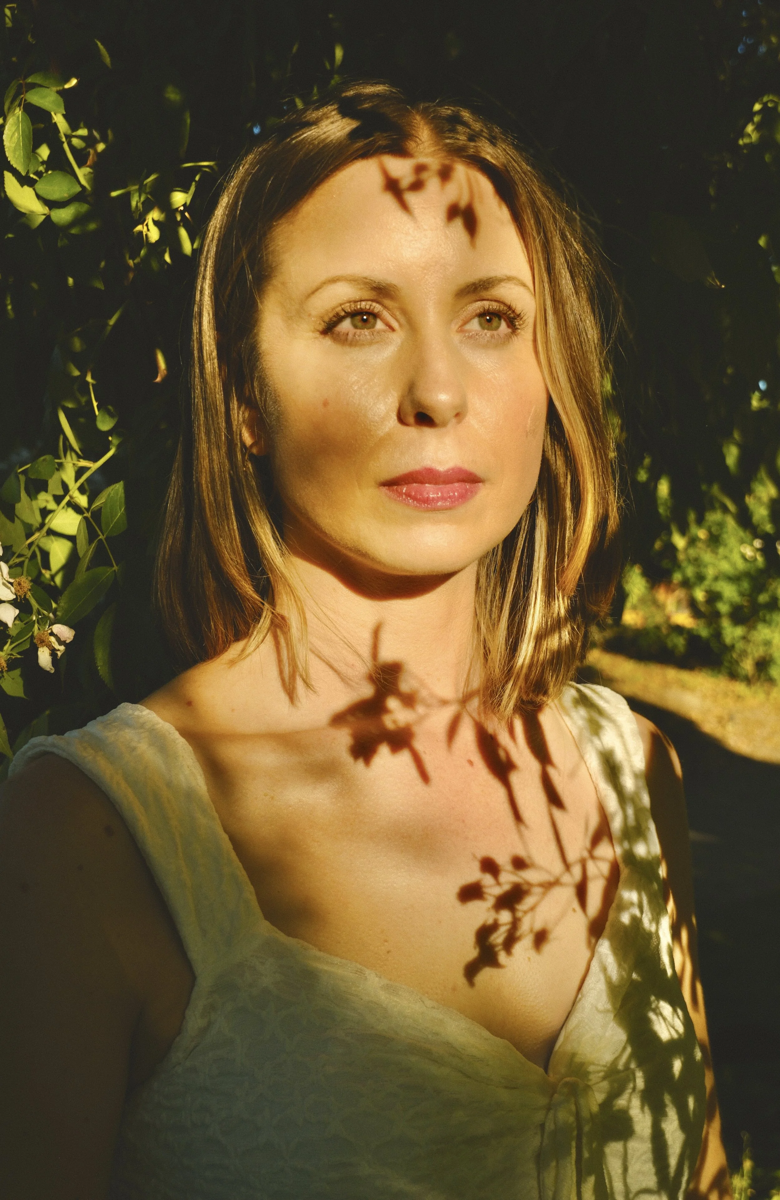 A woman with shoulder-length hair and green eyes standing outdoors among greenery, with sunlight casting shadows of leaves on her face and shoulders.