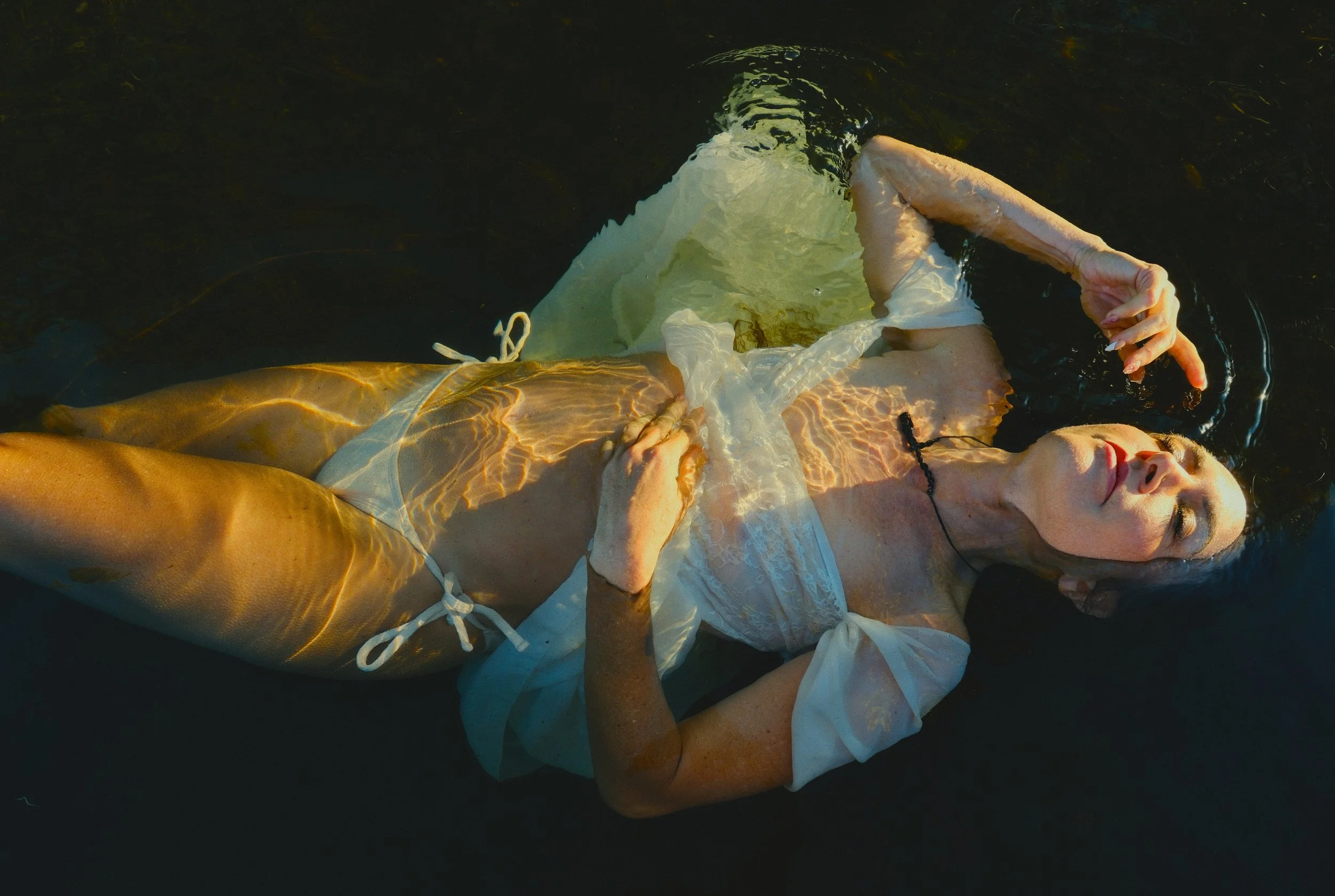 A woman lying in water, wearing a white dress, with her eyes closed and head tilted to the side.