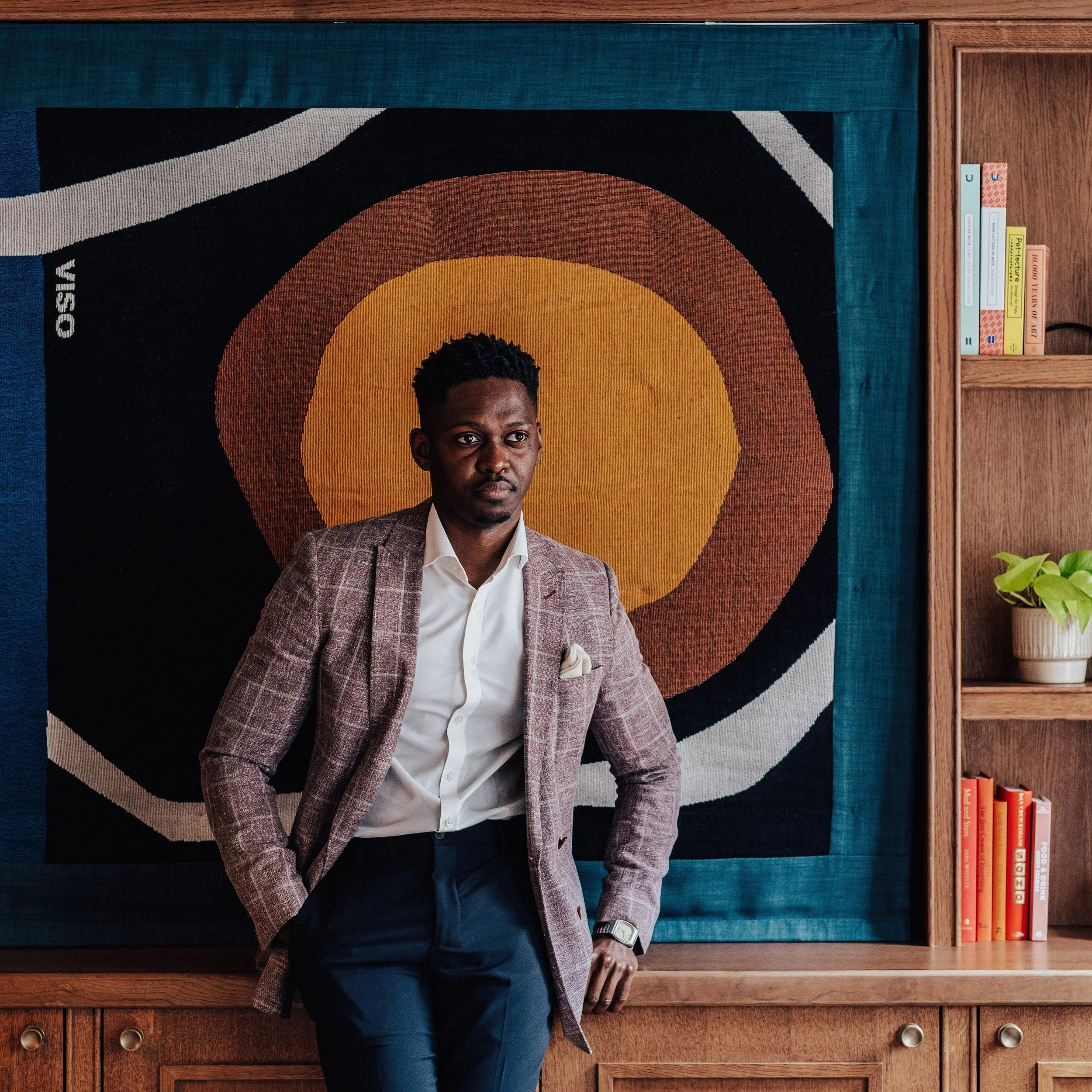 A man in a plaid blazer and white shirt posing in front of an abstract art piece with concentric shapes in orange, yellow, and brown behind him, on a wooden wall with a bookshelf and potted plant nearby.