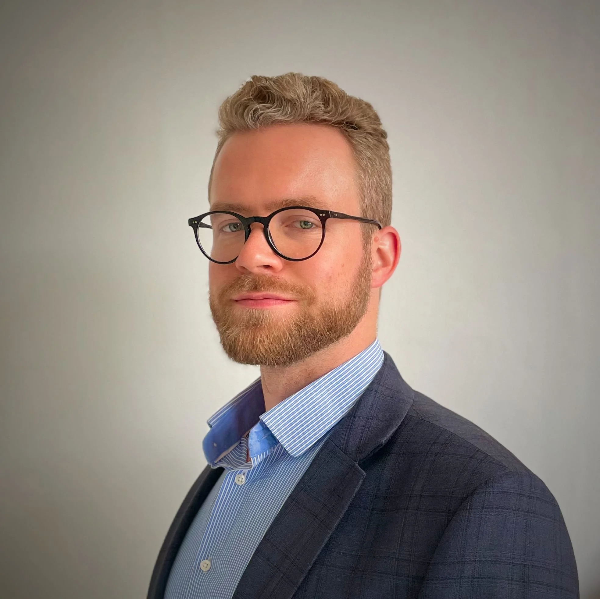 A man with glasses and a beard dressed in a business suit against a plain background.