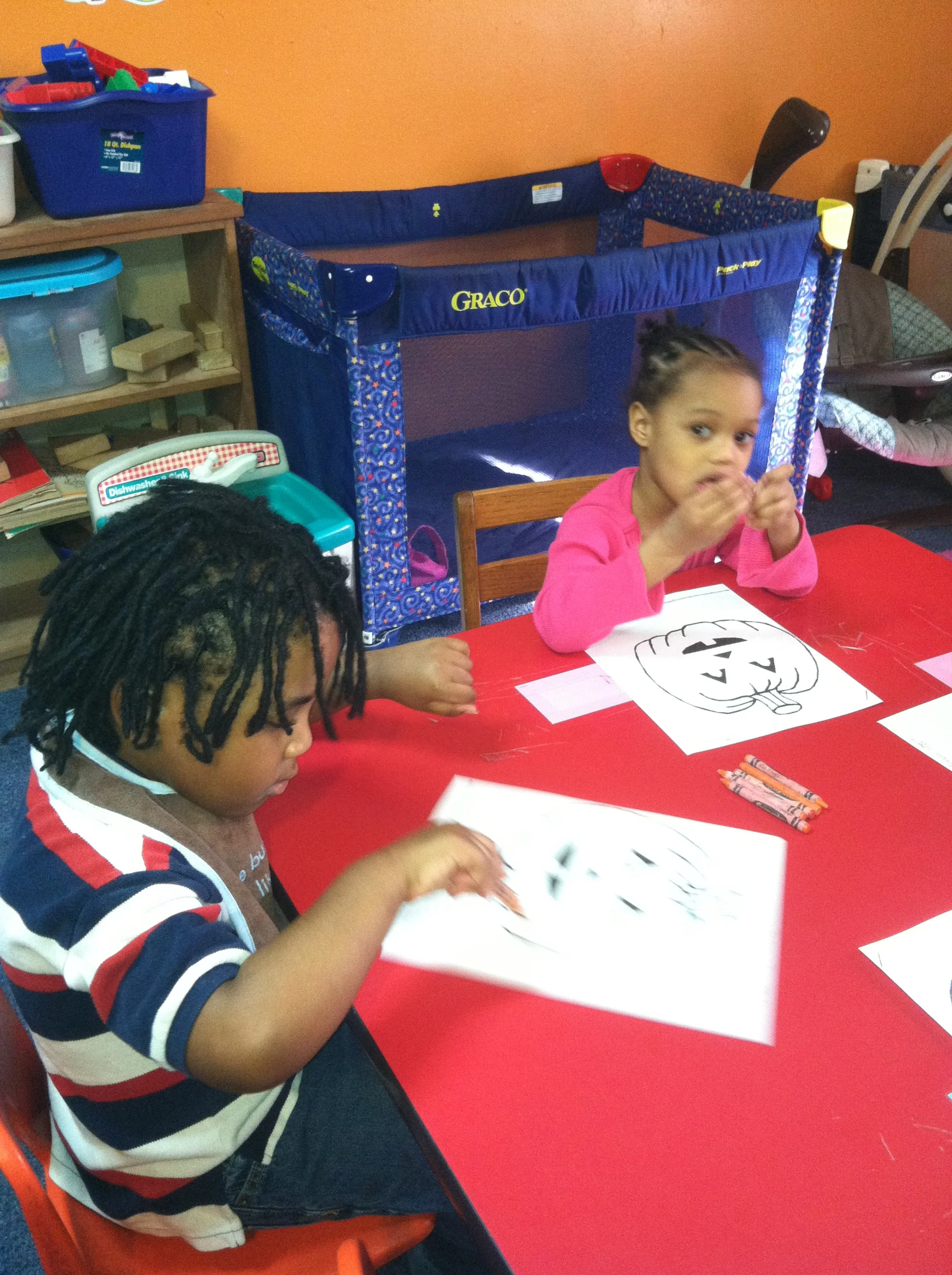 Two children sitting at a red table, coloring Halloween-themed pictures of pumpkins, with a playpen and toys in the background.