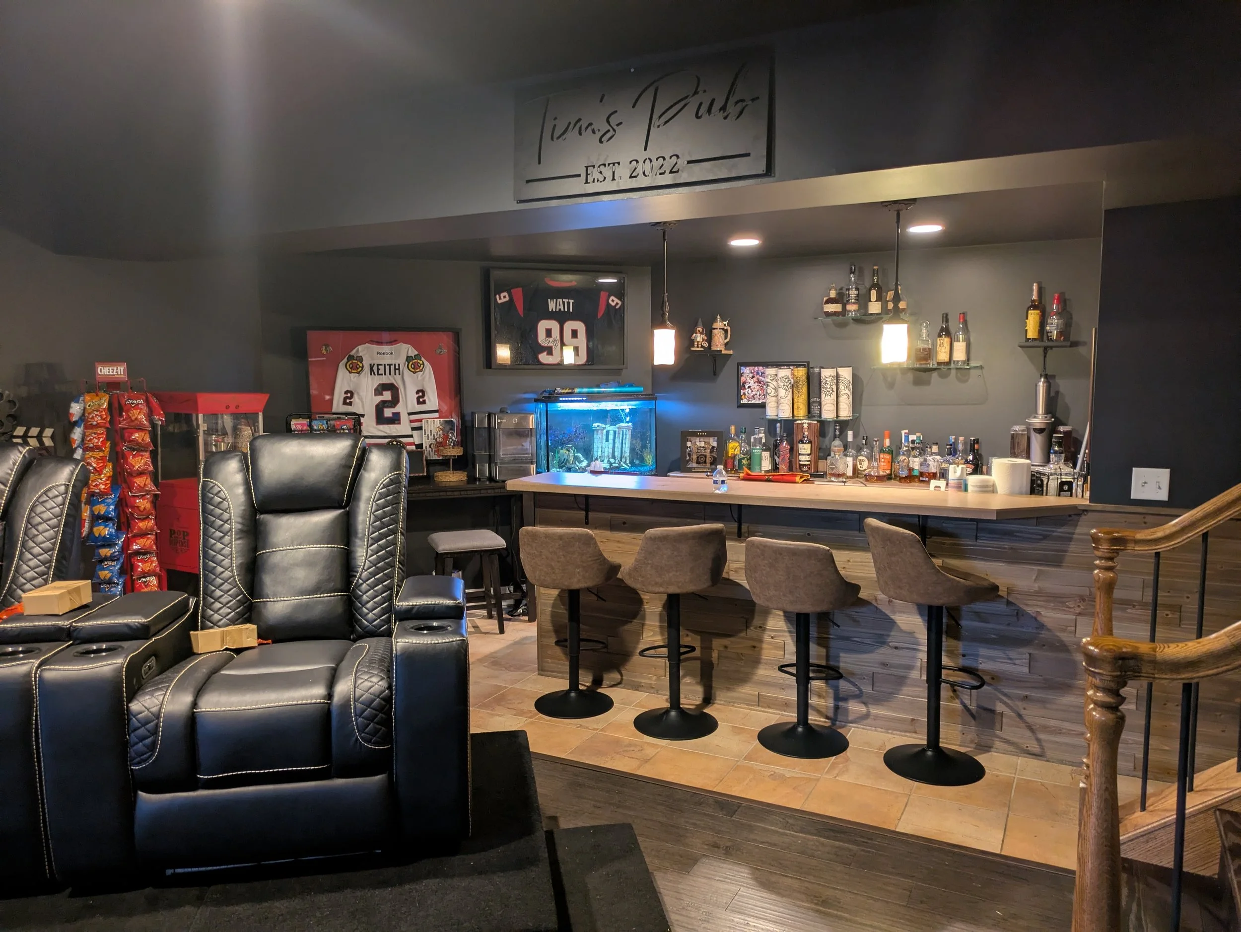 A cozy home bar with a dark wall, a wooden bar counter, four bar stools, various bottles of alcohol, a fish tank, framed hockey jerseys, and a popcorn machine in the corner.