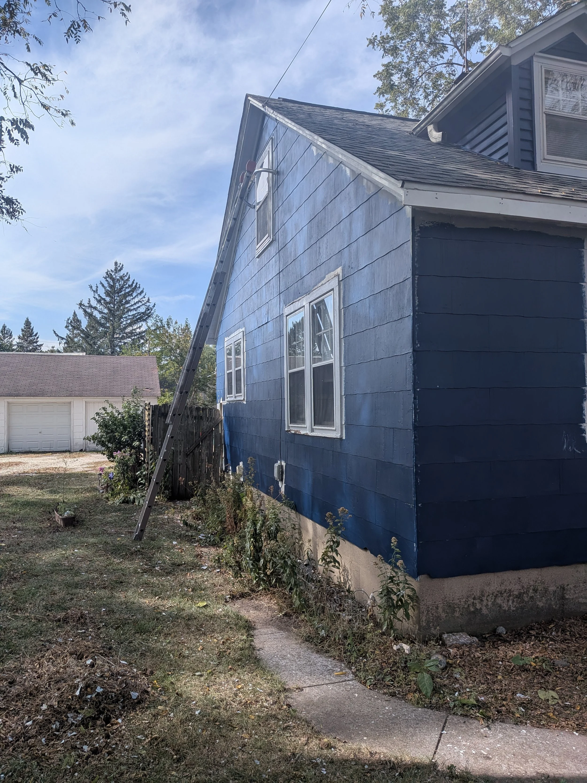 Side of a blue house with white trim, with a ladder leaning against the roof, and a sidewalk leading to the yard with some plants and a fence.