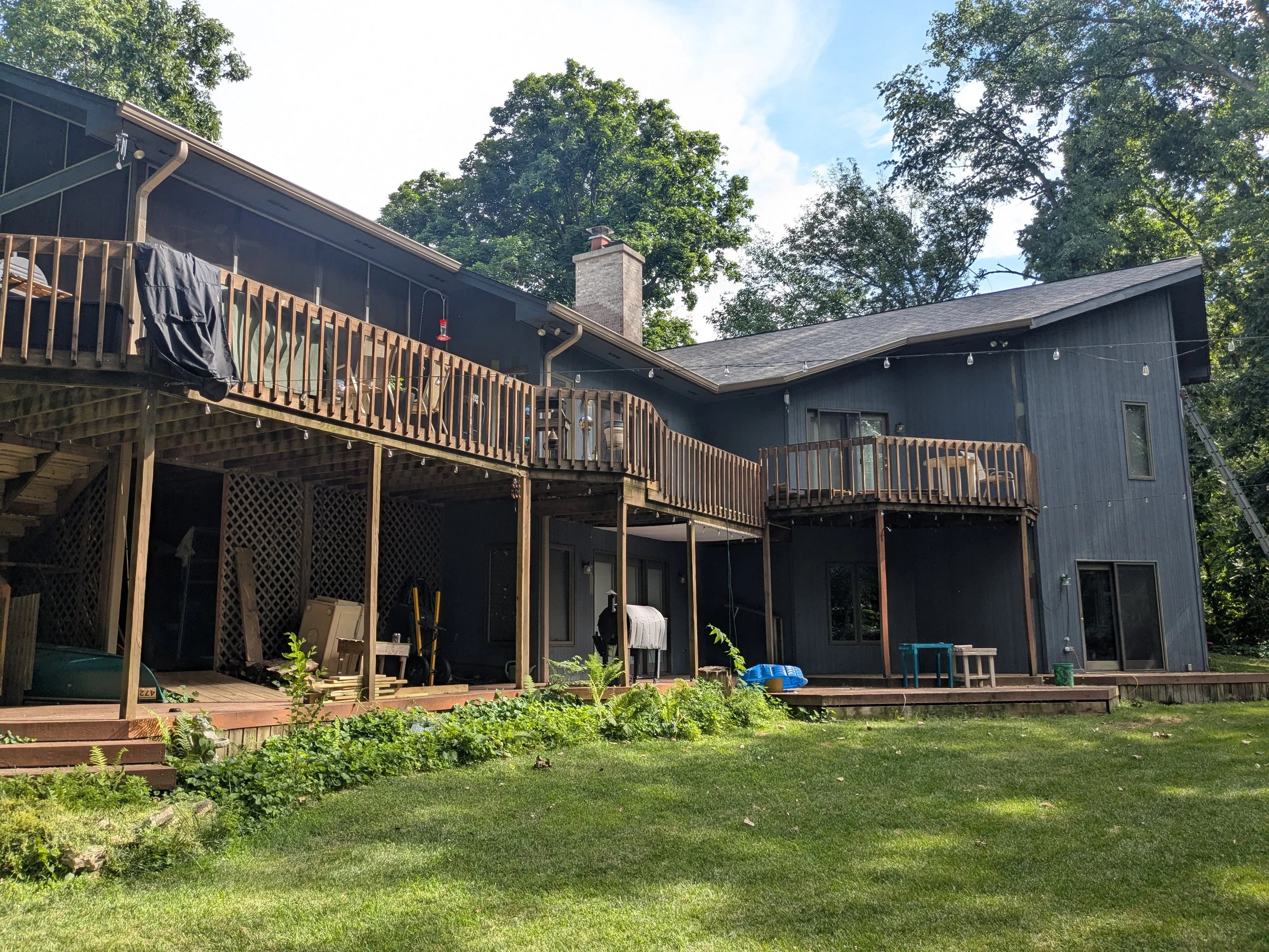 Rear view of a two-story house with a wooden deck, surrounded by a grassy yard and tall trees in the background. The house is painted dark blue with large windows and a chimney.